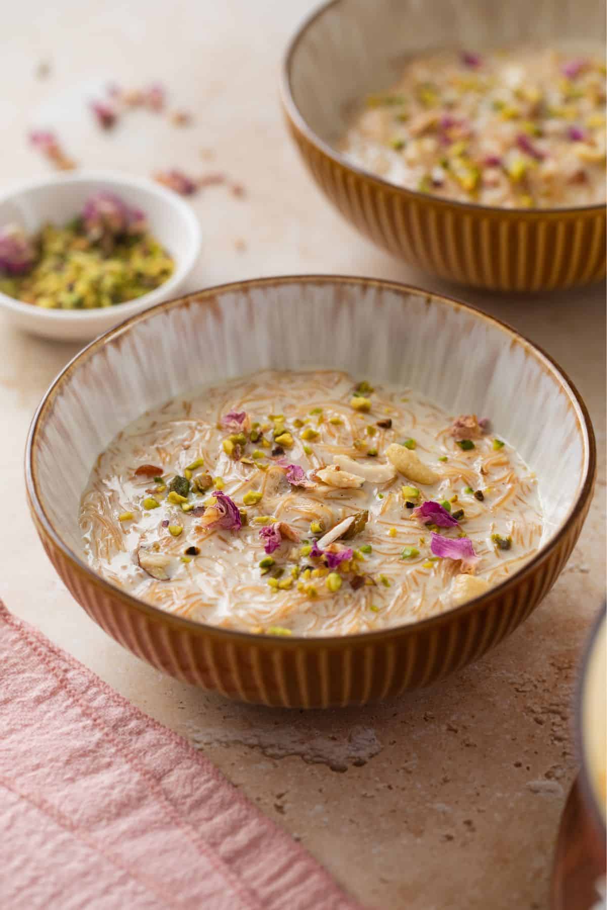 Indian Seviyan Recipe (Creamy Vermicelli Kheer ) 3 Side image of a bowl of A bowl of creamy seviyan kheer garnished with chopped nuts and dried rose petals, placed on a light surface with a pink cloth.