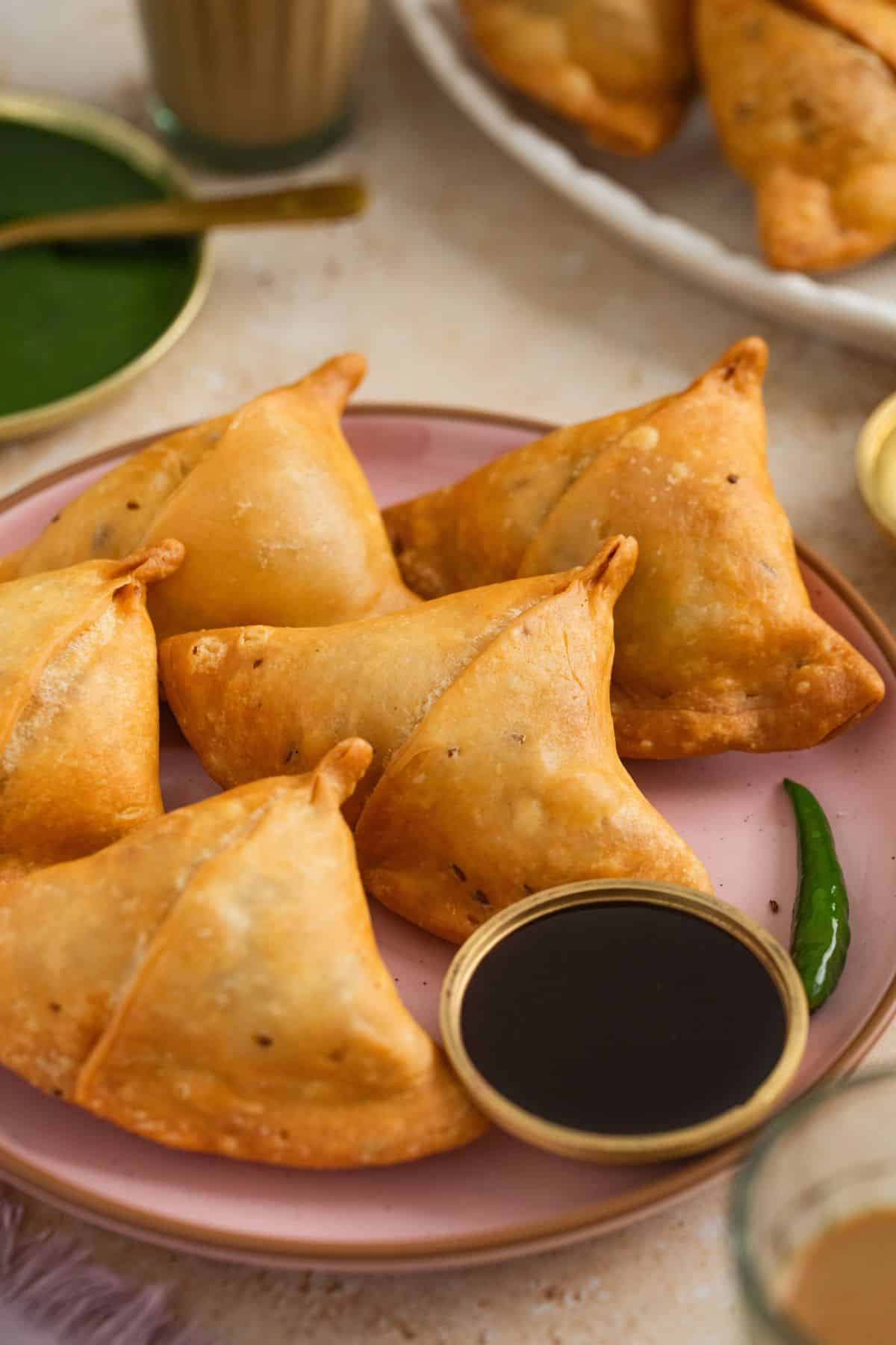 Classic Punjabi Samosa Recipe (Crispy & Flaky) 3 A pink plate with five golden-brown samosas, served with a small bowl of tamarind and green chutney.Other dishes and a glass of chai are visible in the background.