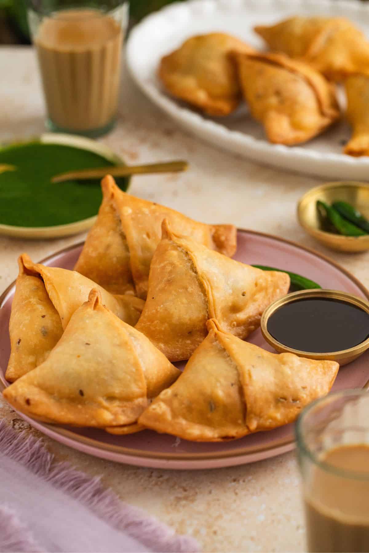 Classic Punjabi Samosa Recipe (Crispy & Flaky) 39 A table scenece with two plates of samosa, chai glasses, fried green chillies and chutneys.