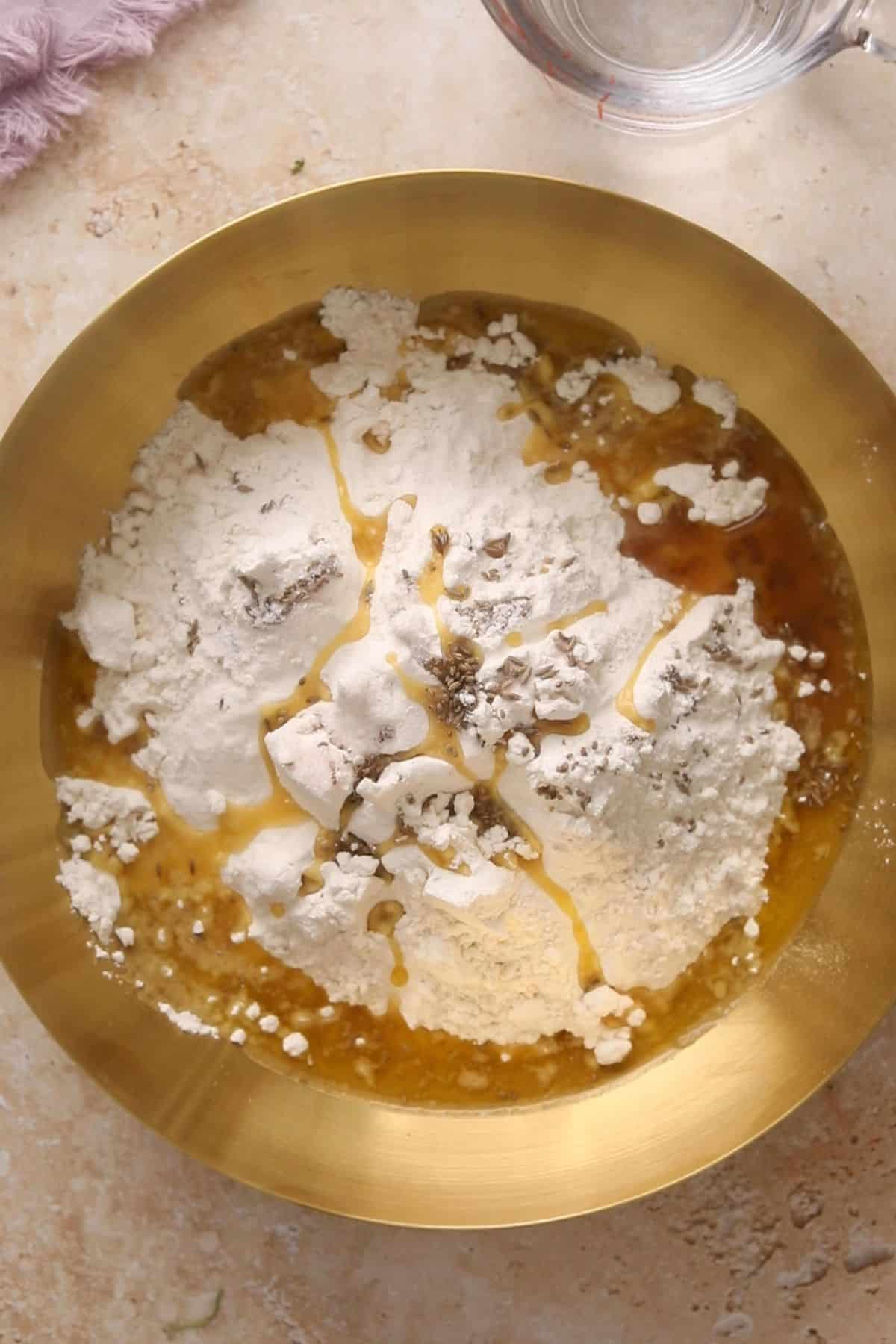 A gold bowl with flour, oil, water, and spices being mixed together on a light countertop. A measuring cup with water is partially visible nearby.