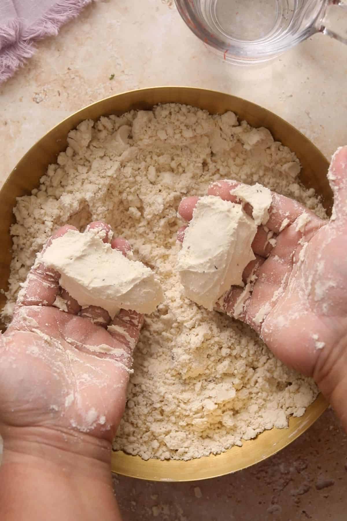 Hands holding and crumbling flour mixture over a bowl, with some water and a lavender cloth visible in the background on a light countertop.