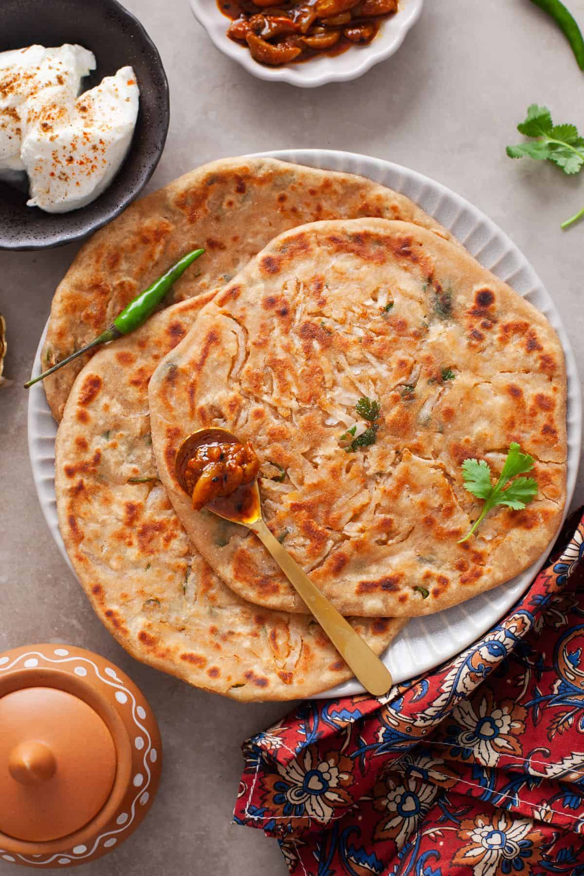 Punjabi Mooli Paratha (Stuffed Radish Paratha) 3 Mooli paratha served on a white plate with garlic pickle and yogurt on side.