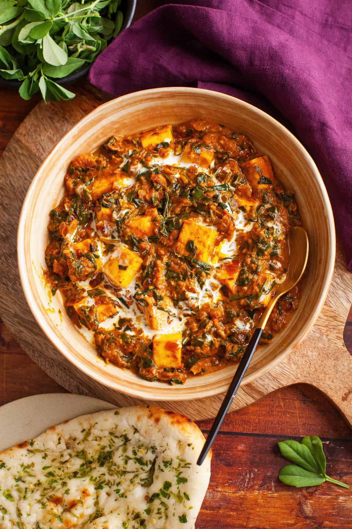 Methi paneer is served in a beige bowl with a gold and black spoon. A chili naan and fresh methi is also visible in the frame.