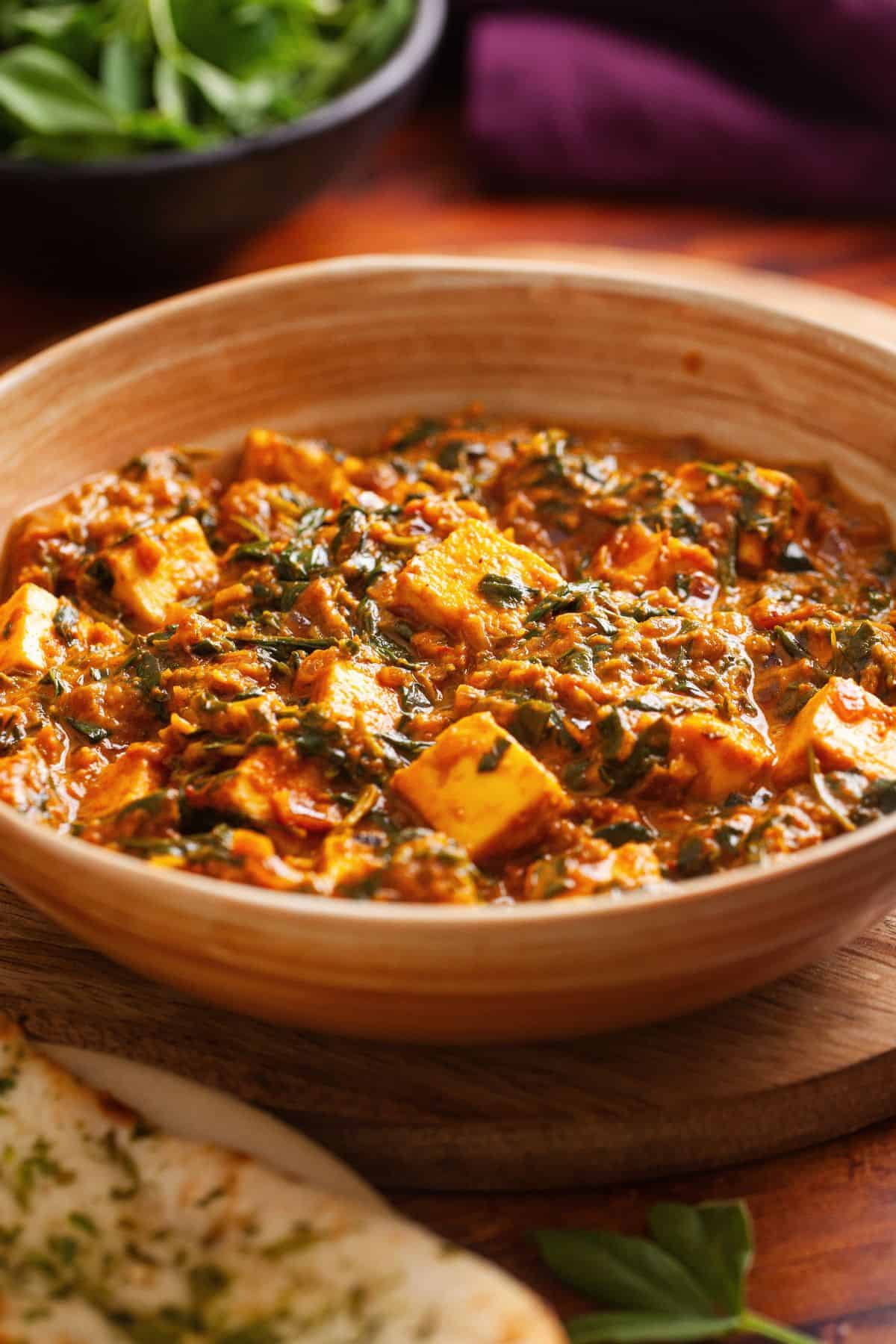 Side view of paneer methi curry served in a shallow beige bowl with purple cloth and methi in background.