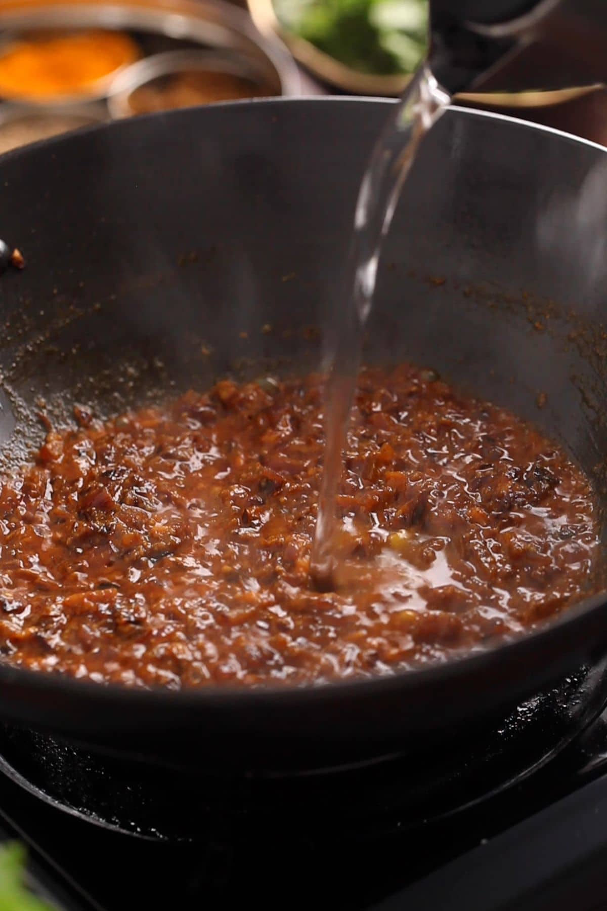 Adding hot water to the onions and spices while frying the masala.