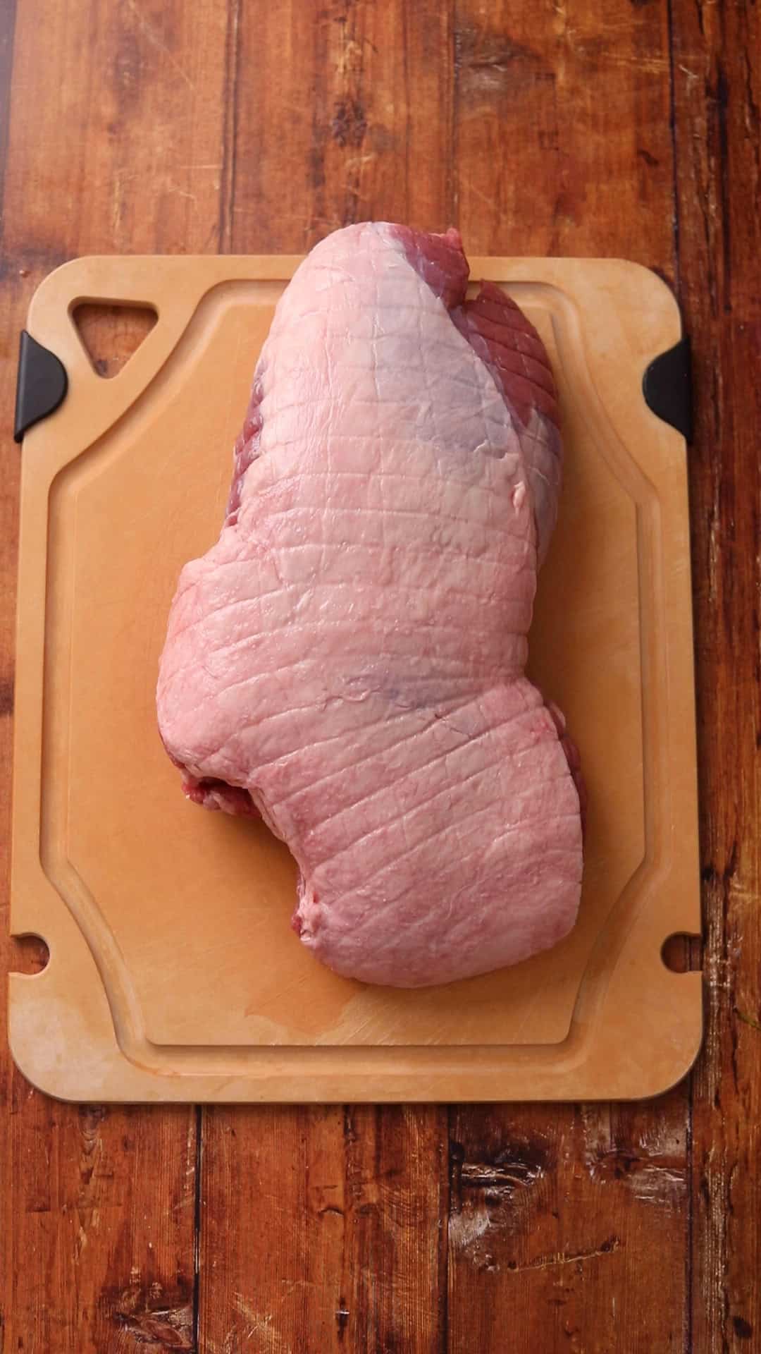 Boneless leg of lamb rests on a tan plastic cutting board. 