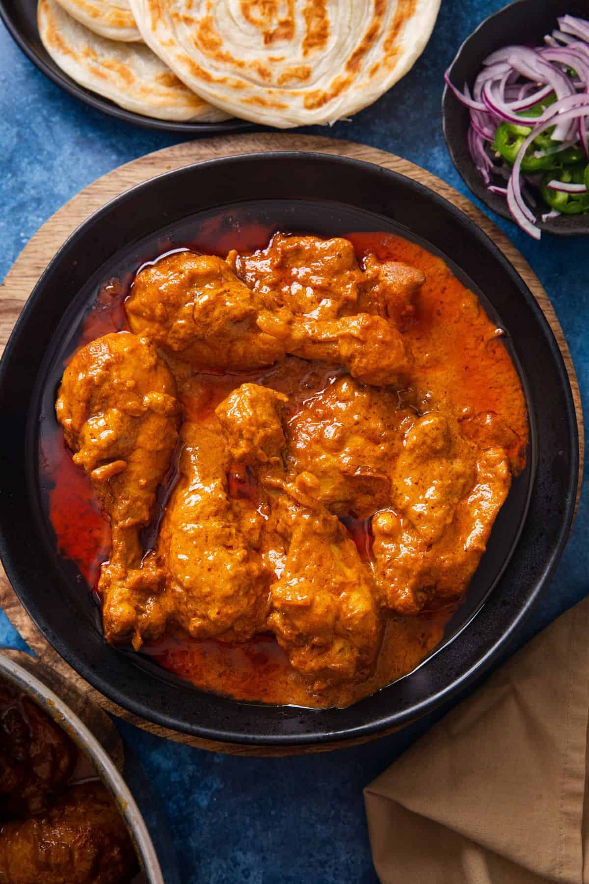 Delhi Chicken Korma is served in a wide and shallow black bowl with sides such as flaky flatbreads, sliced onions and green chillies. Everythung is placed on a blue background.