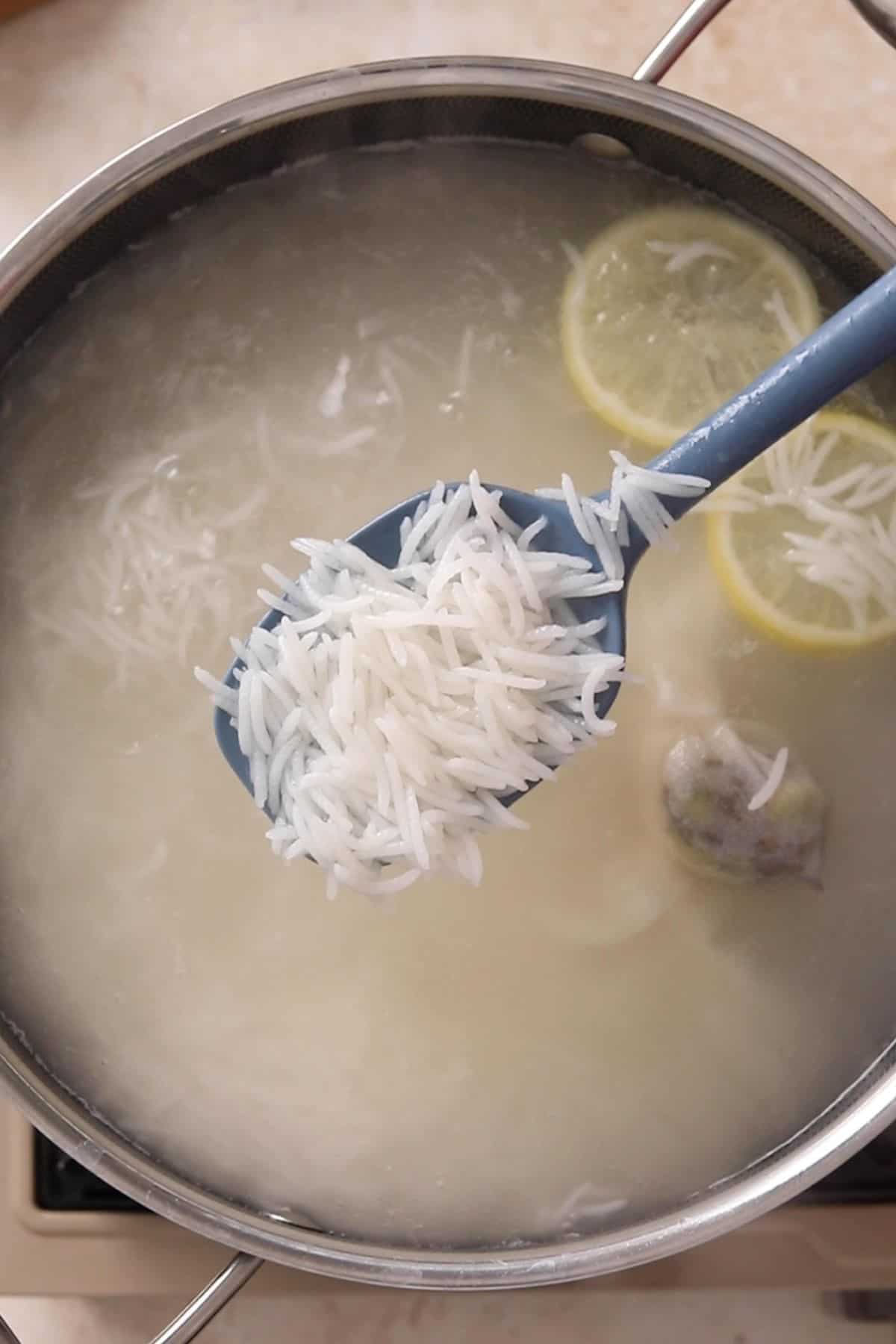 A close-up of a spoon holding cooked white rice above a pot of boiling water with lemon slices and a spice bag floating in it.