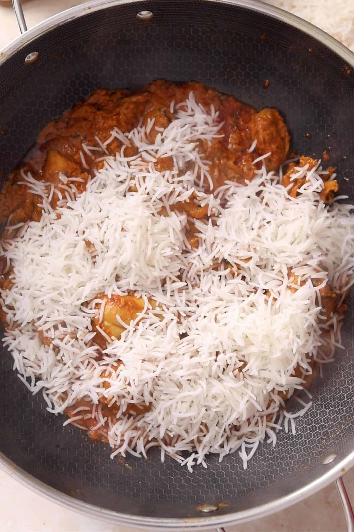 A pot filled with cooked basmati rice being added to a tomato-based curry with chunks of vegetables or meat, ready to be mixed together.