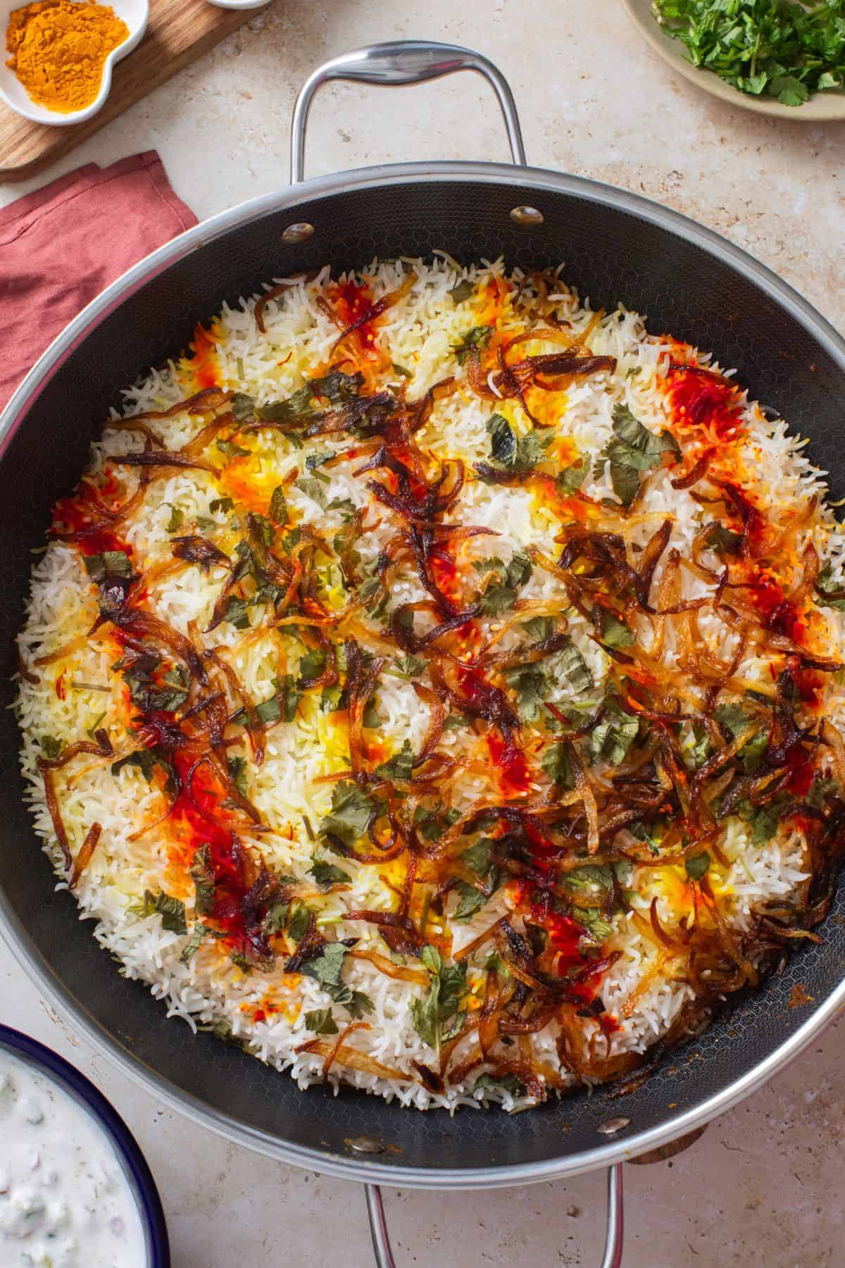 A pan of biryani topped with caramelized onions, herbs, and saffron, showing vibrant colors of yellow, orange, and green over fluffy white rice. Dishes with yogurt and chopped herbs are nearby on a light countertop.