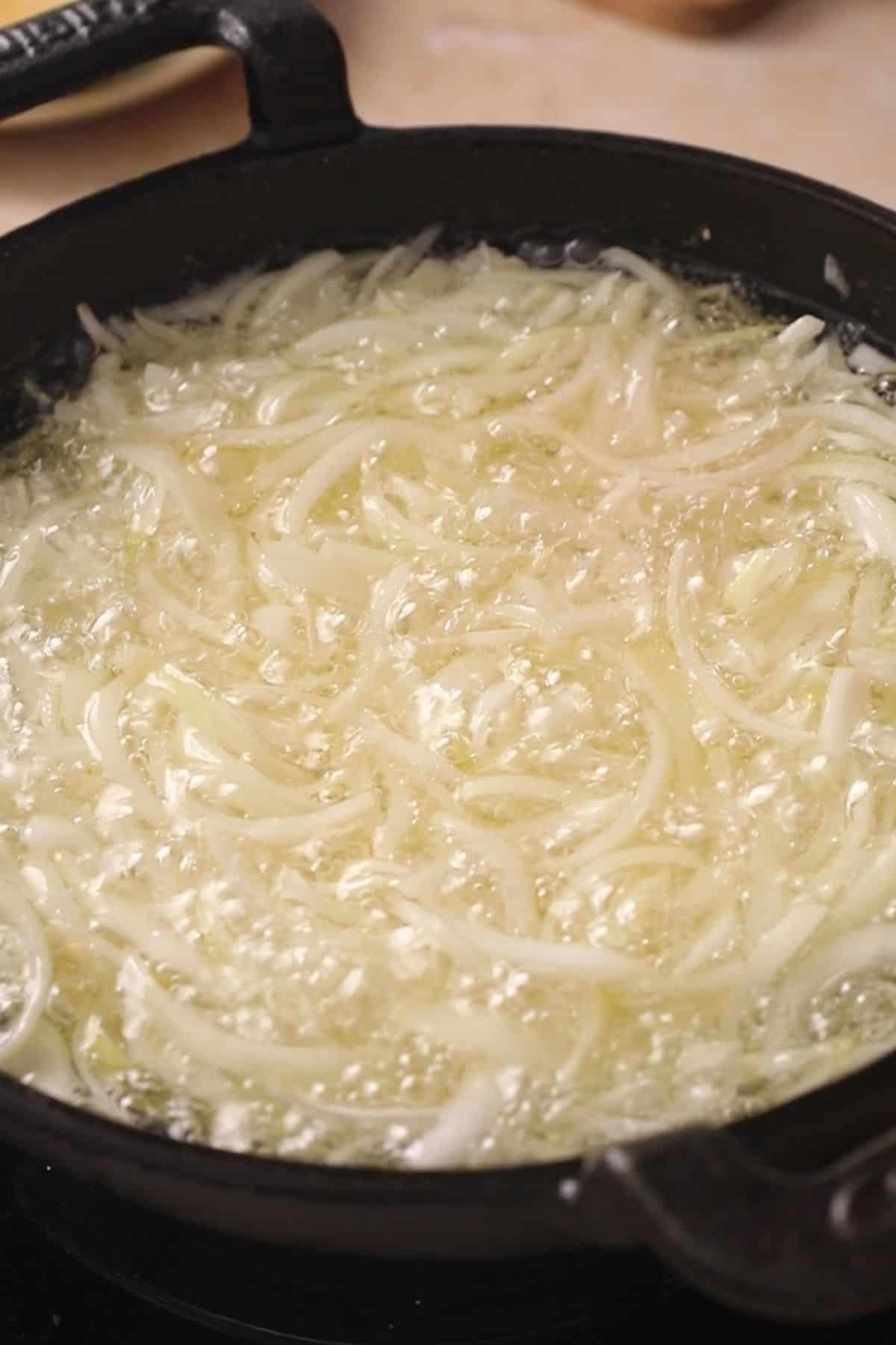 Thinly sliced onions frying in hot oil in a black cast iron skillet, with bubbles forming around the onions as they cook.