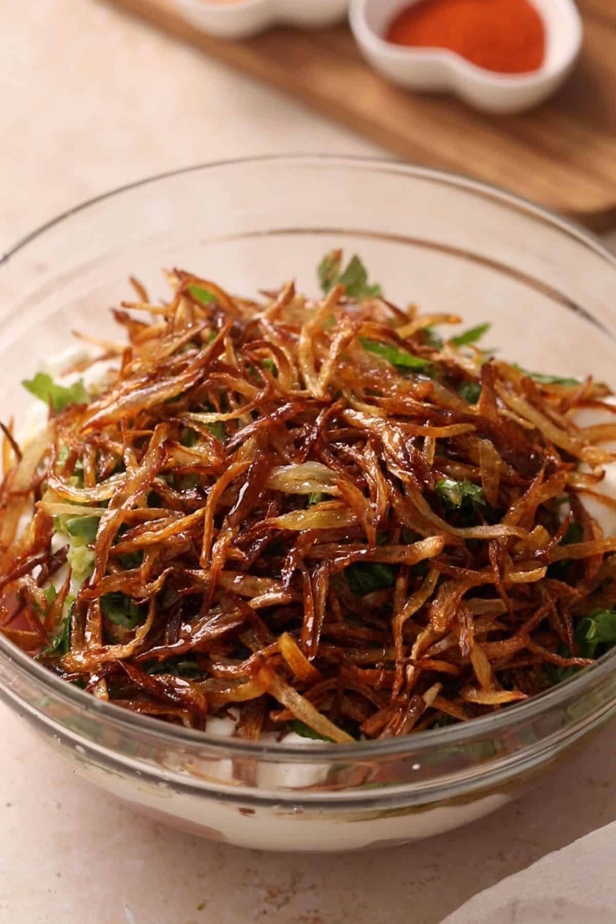 A glass bowl containing raw chicken pieces topped with yogurt, grated ginger, and piled spices including turmeric, cumin, coriander, birista and red chili powder, ready for marination.