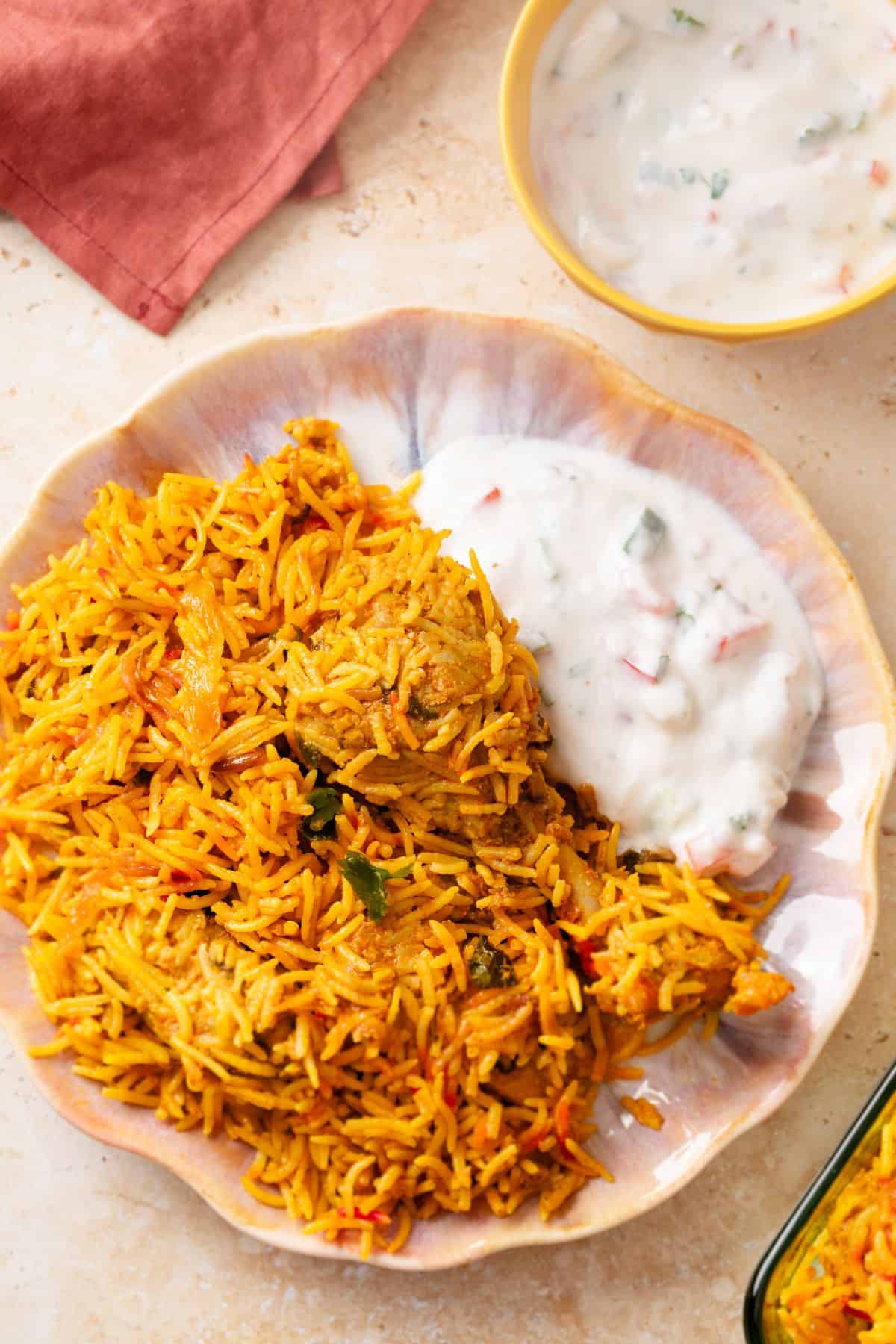 A plate of biryani rice with pieces of chicken, served alongside a portion of creamy yogurt raita. The background includes a yellow bowl of raita and a red napkin.