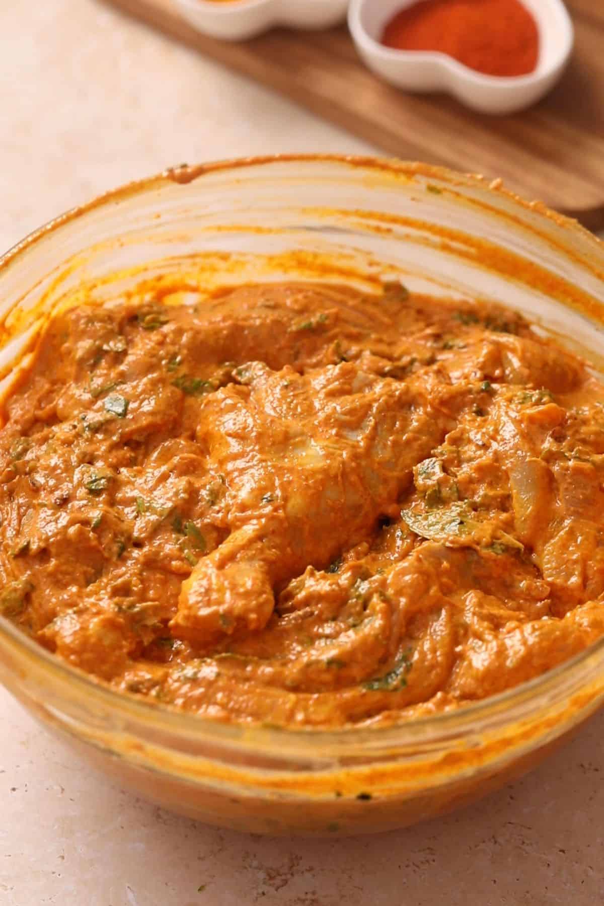 A glass bowl filled with chicken pieces marinating in an orange, creamy, and spiced marinade with visible herbs.