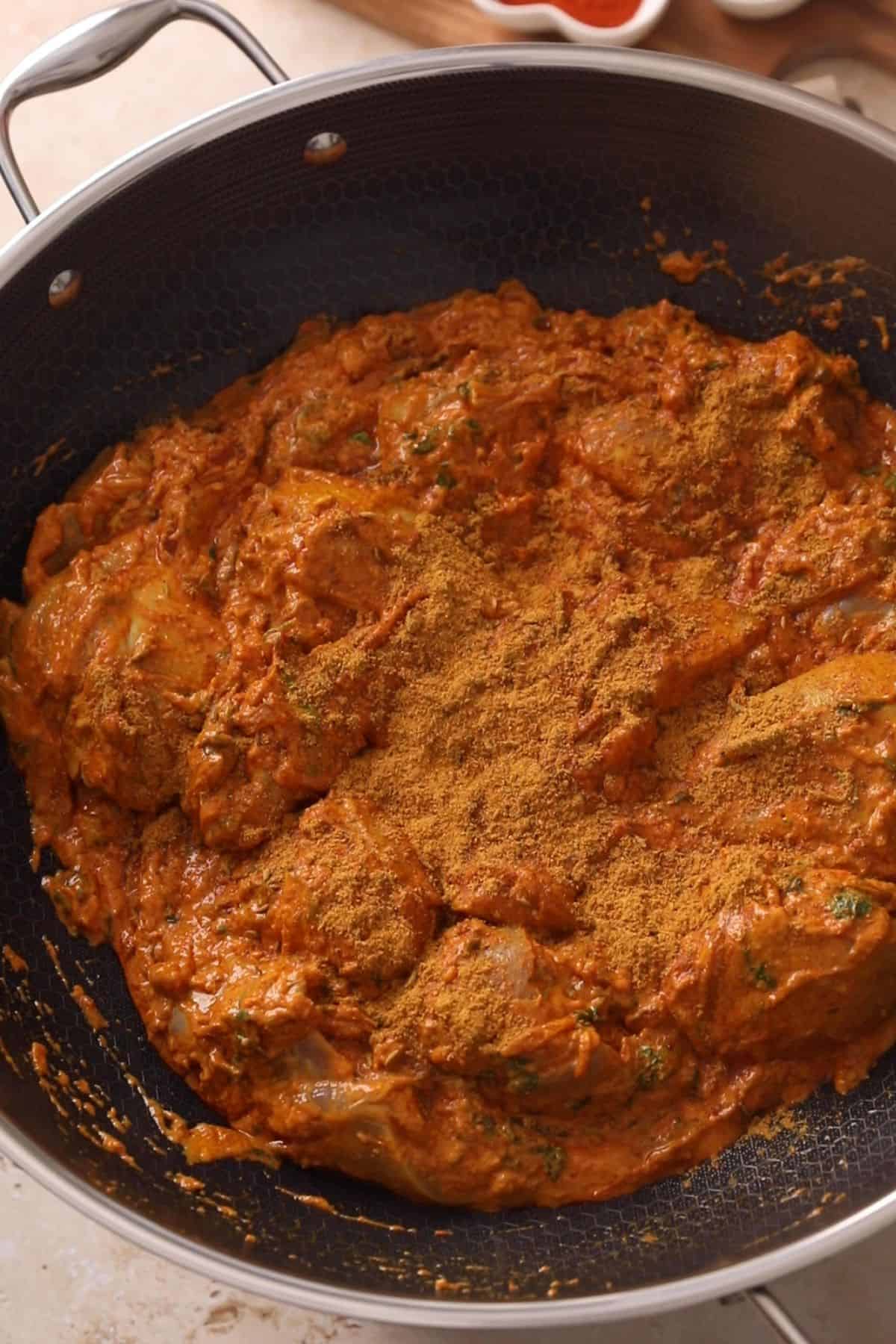 Marinated chicken pieces coated in a thick, orange-brown spiced yogurt marinade with a generous sprinkle of dry spices on top, cooking in a large pot.