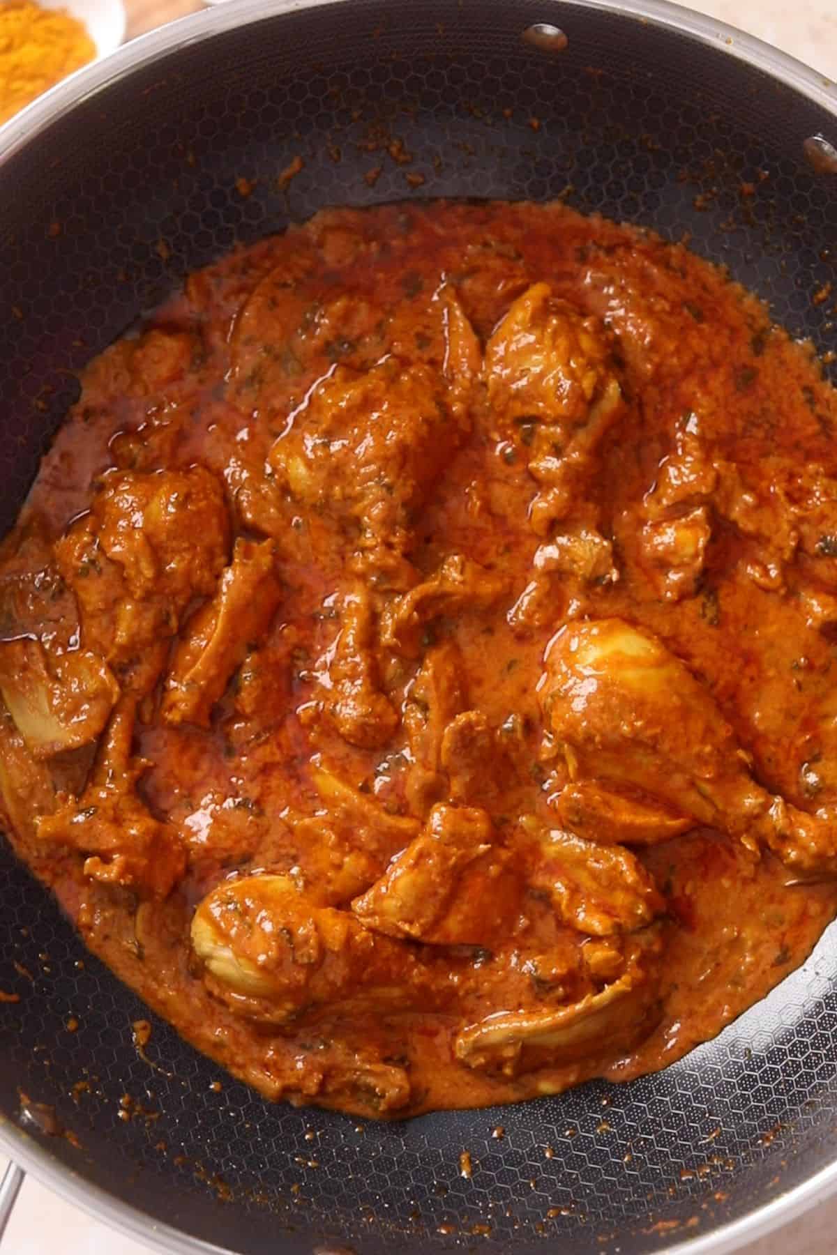 Chicken pieces simmer in a rich, red-brown curry sauce inside a deep, pot, with visible spices and oil on the surface, creating a hearty and flavorful chicken base for biryani.