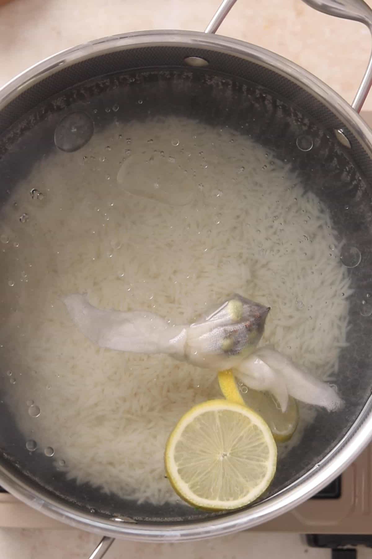 A stainless steel pot filled with water, uncooked white rice, a tied bundle of herbs or spices, and two slices of lemon floating on top. The pot is on a stovetop.