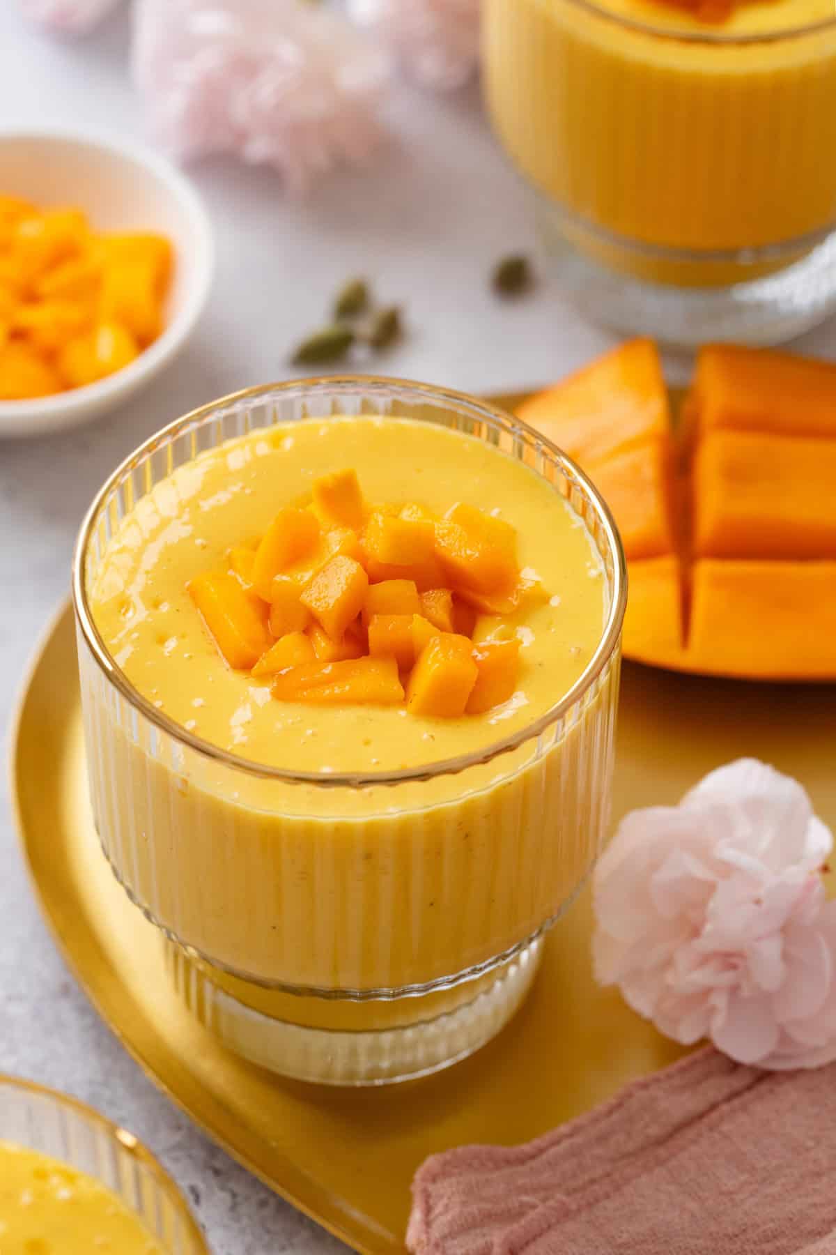 A glass of creamy mango lassi garnished with diced mango pieces sits on a gold tray, with sliced mango and pink flowers nearby. A bowl of extra mango cubes is in the background.
