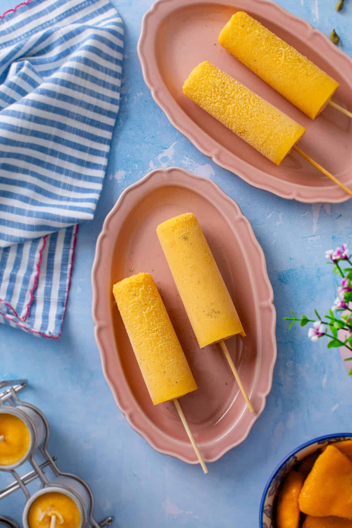 Three yellow mango kulfi on oval pink plates are arranged on a light blue surface, with a striped blue and white cloth nearby and some flowers and metal molds partly visible.