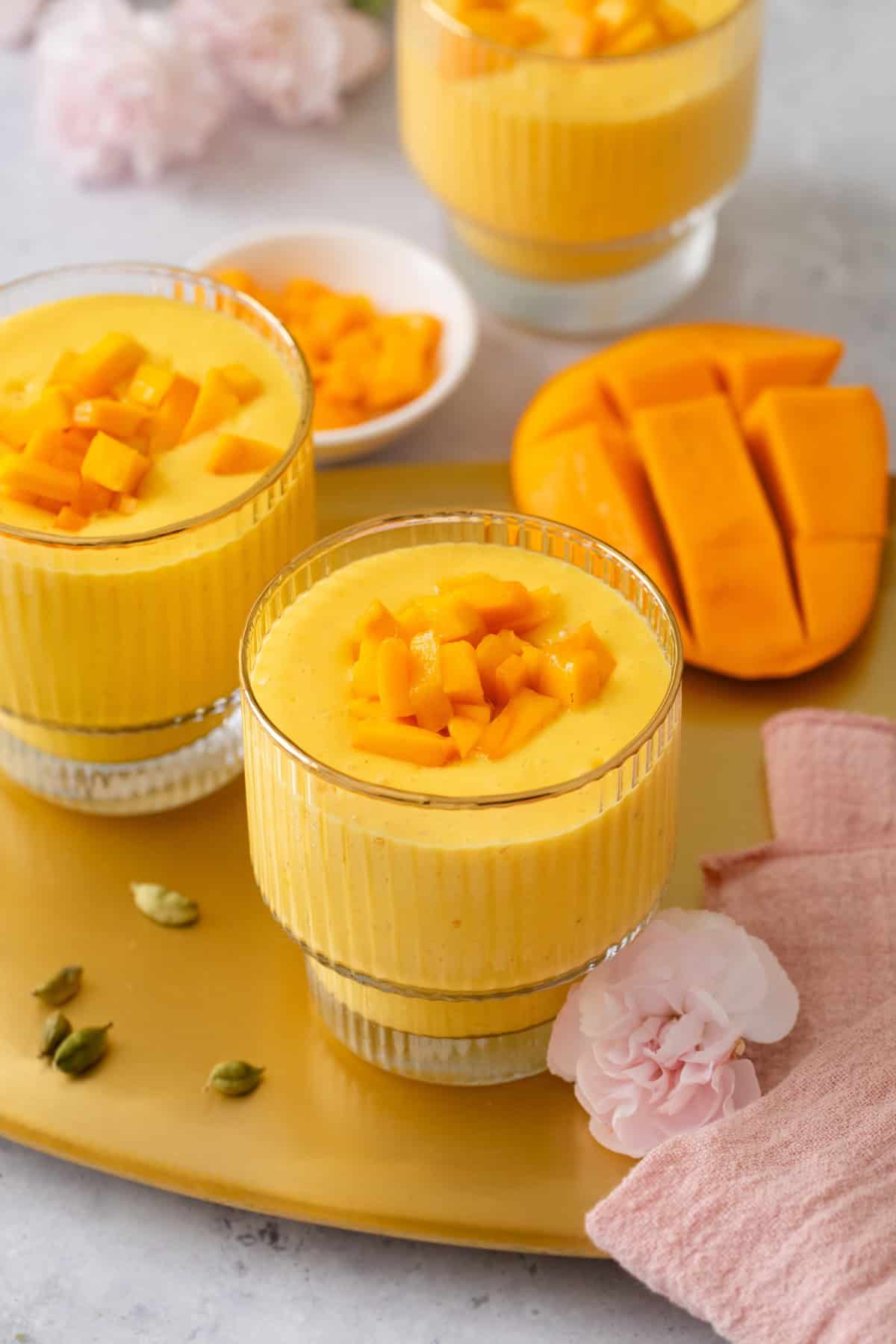 Three glasses of mango lassi topped with diced mango, placed on a gold tray with sliced mango, cardamom pods, pink flowers, and a pink napkin in the background.