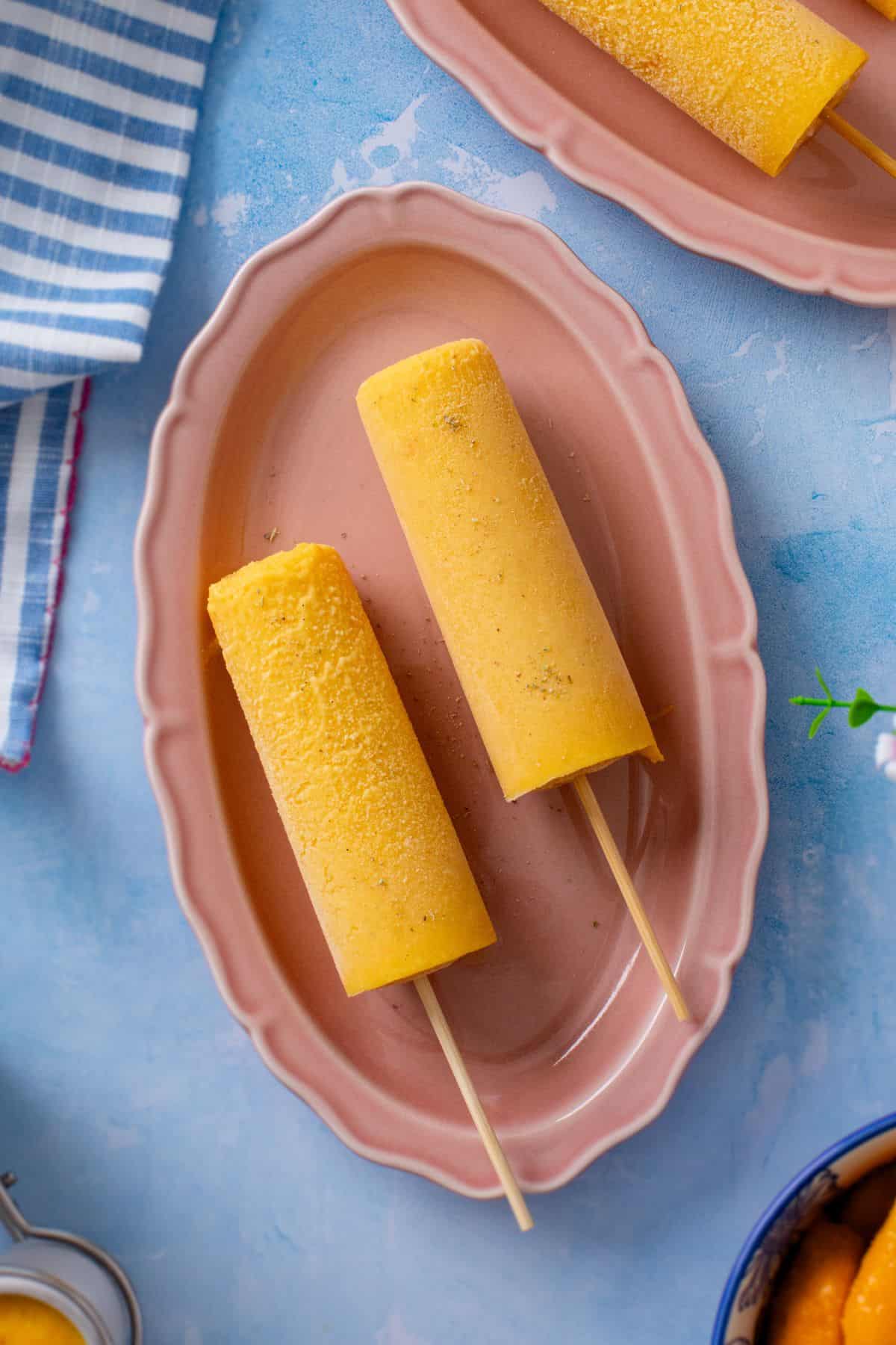 Two mango kulfi are placed in a oval pink plate.