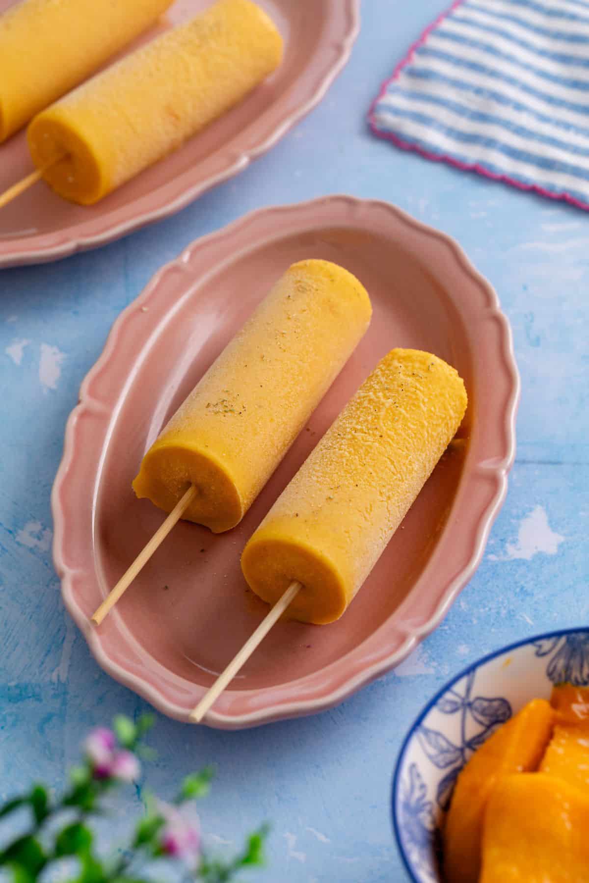 Two mango popsicles on wooden sticks are served on a pink oval plate, with another plate and a bowl of mango pieces nearby, set on a blue background.