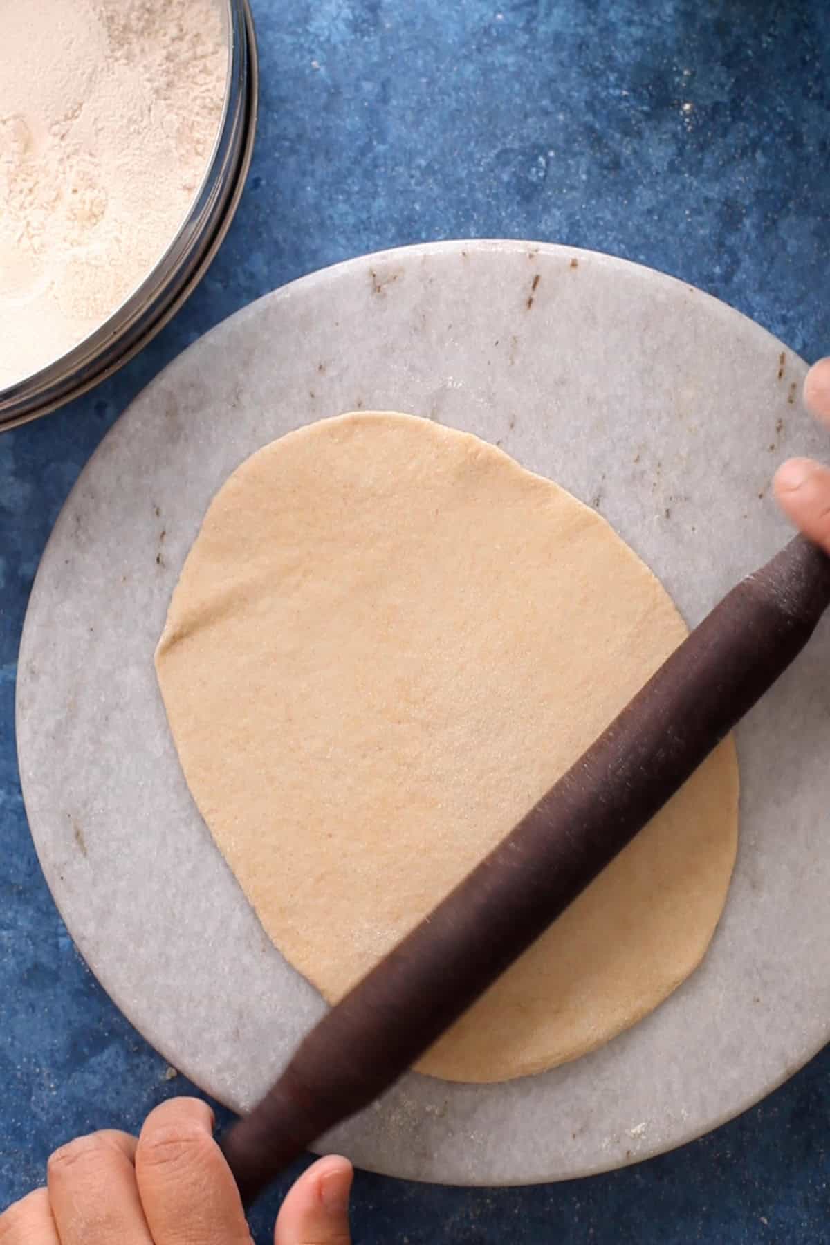 Atta naan is being rolled using a wood rolling pin on a marble stone.