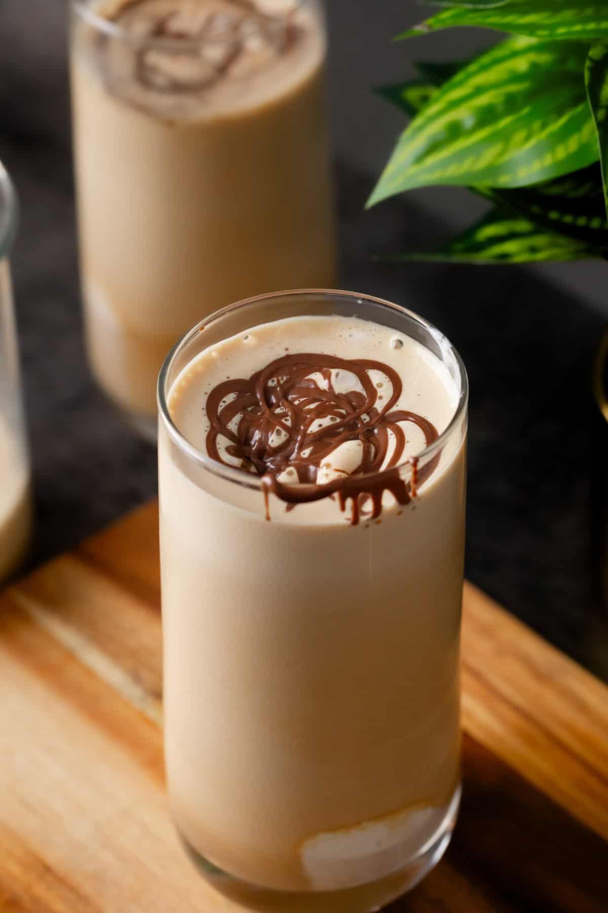 A glass of cold coffee topped with chocolate syrup sits on a wooden surface, with another glass and a green plant visible in the background.