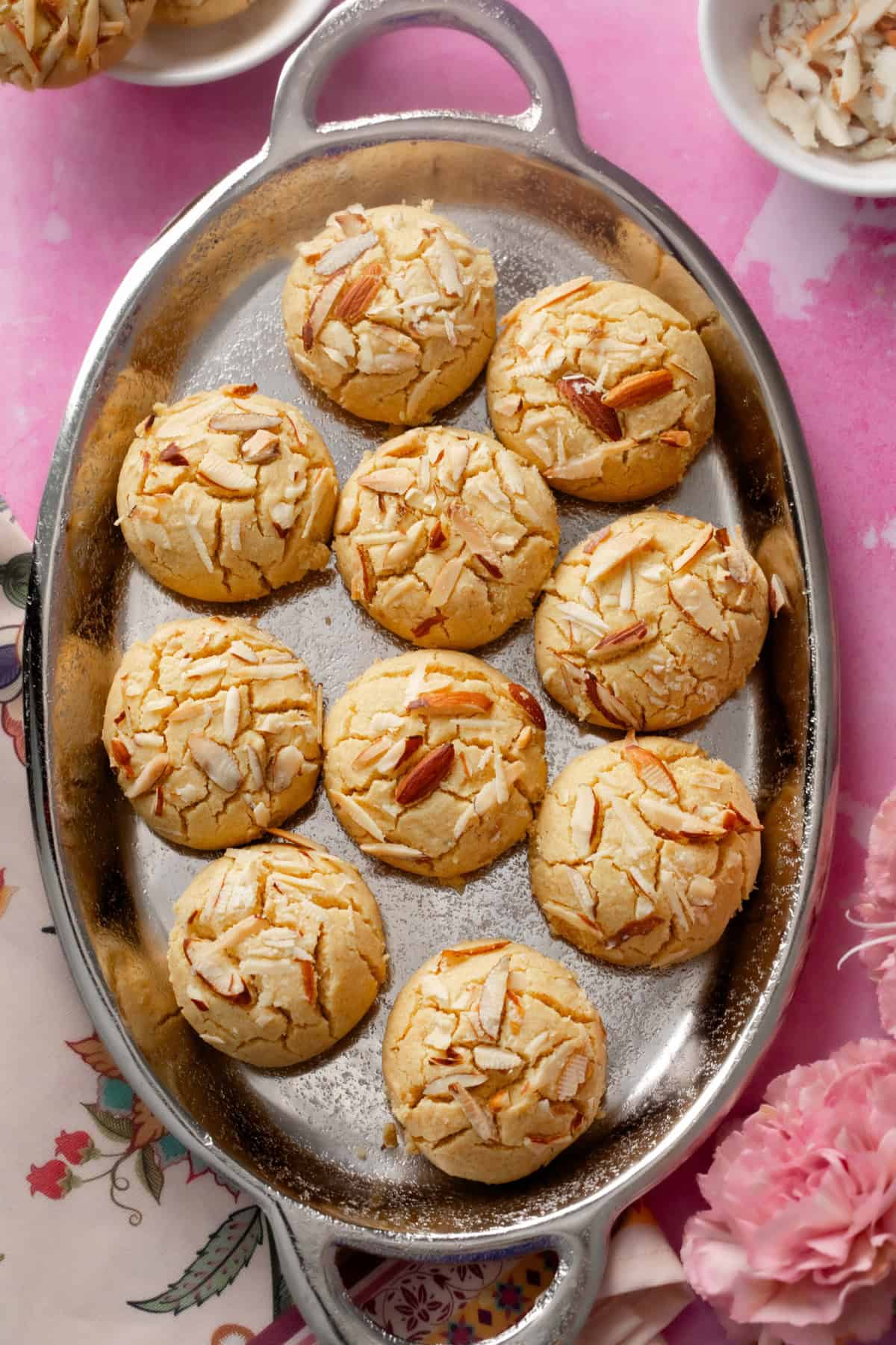 Besan Nankhatai placed on a silver oval platter with sliced almonds & pink flowers on side.