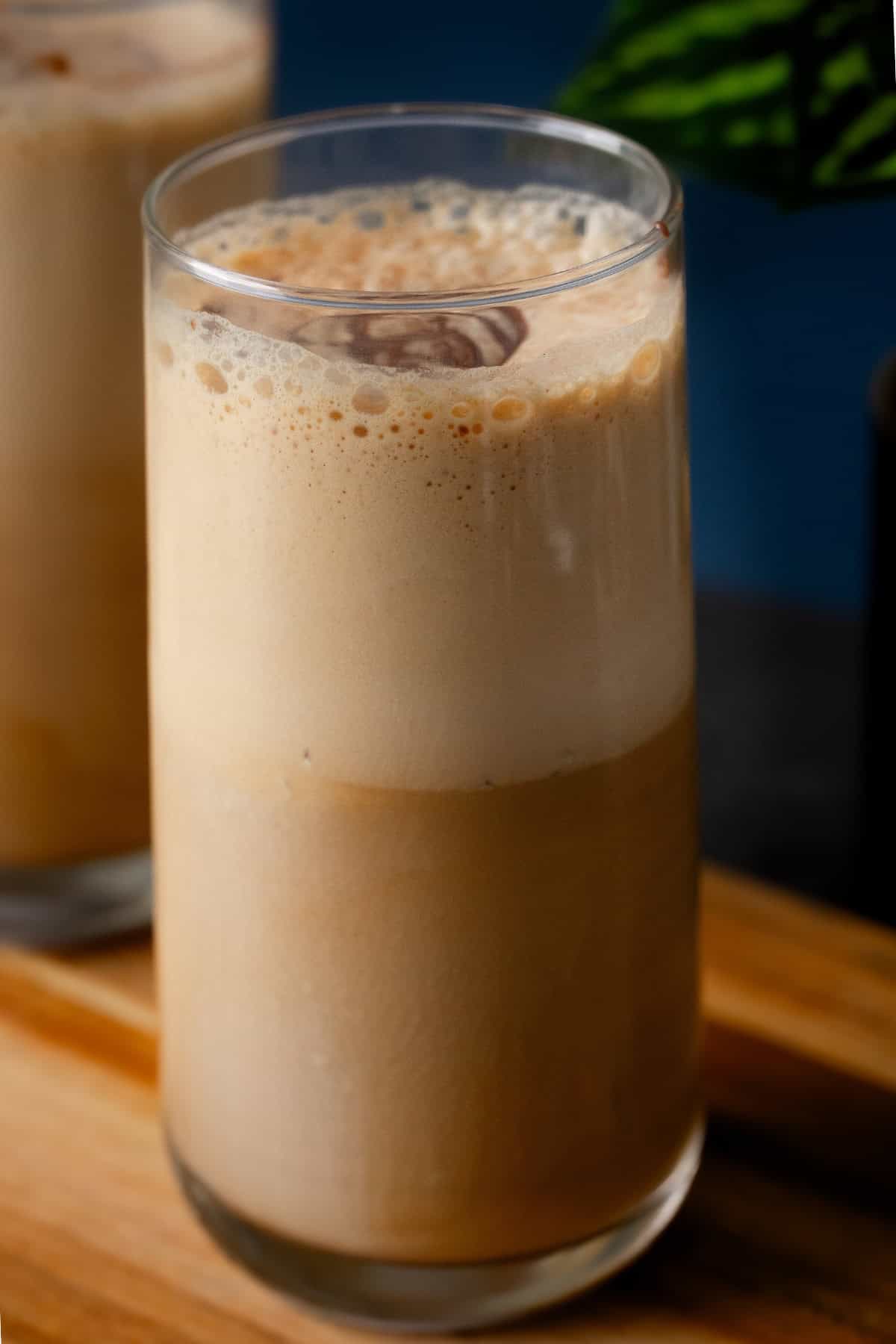 A tall glass filled with creamy cold coffee, with frothy foam, sits on a wooden surface with another glass blurred in the background.