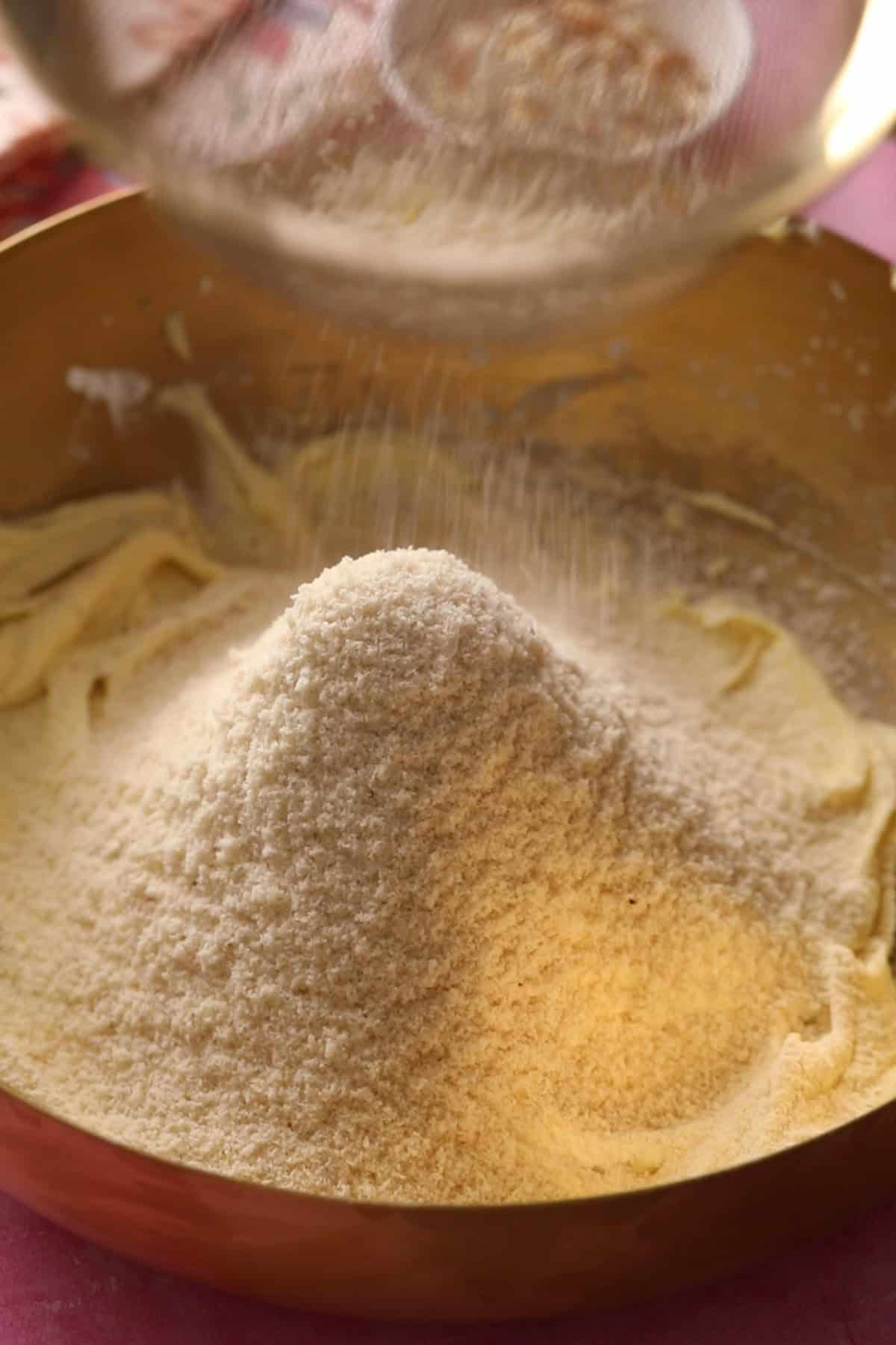 Sifted besan, alomnd flour are added to creamed ghee and sugar in a gold bowl.
