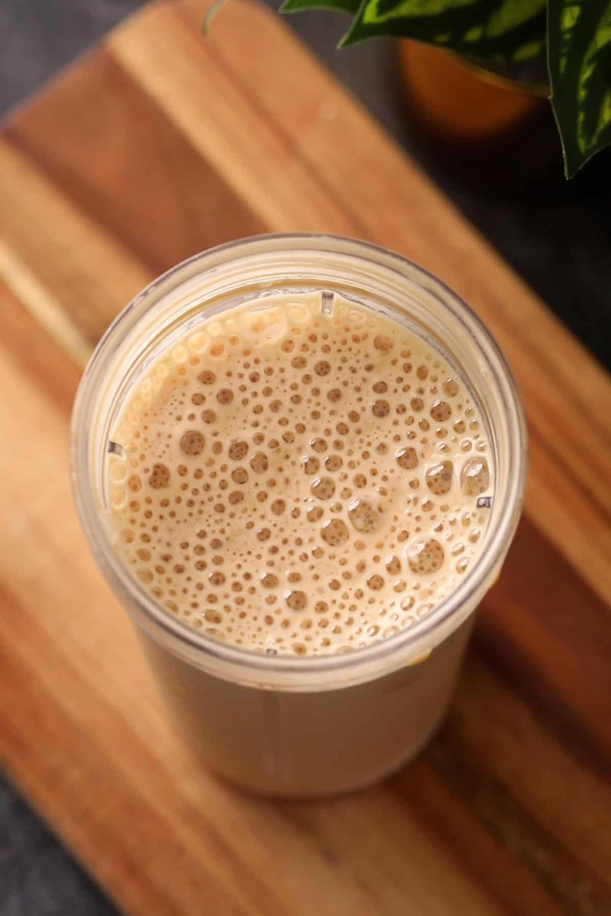 A clear cup filled with a frothy, light brown cold coffee sits on a wooden surface. There is a green plant partially visible in the background.