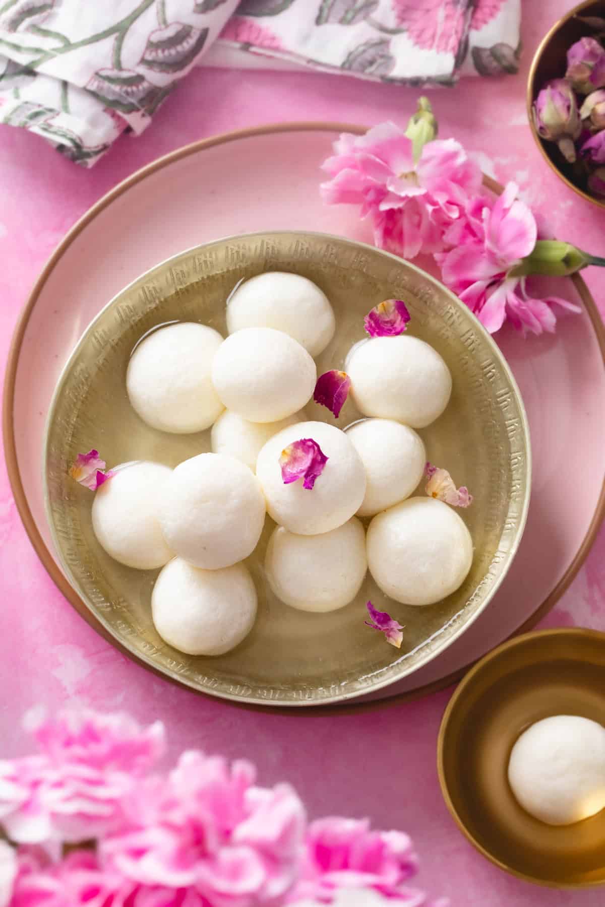 A gold plate with round white rasgullas in syrup, garnished with rose petals, surrounded by pink flowers on a pink surface.