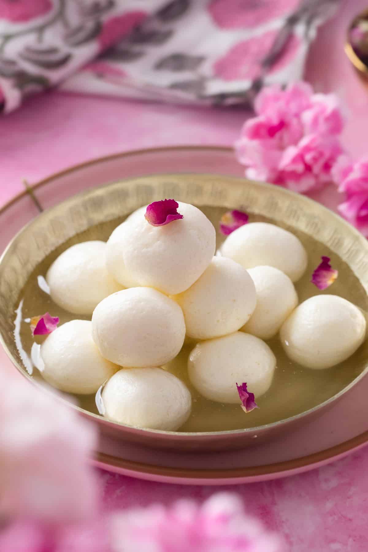 A bowl of round white rasgullas in syrup, garnished with pink rose petals, set on a pink plate with flowers and a patterned cloth in the background.