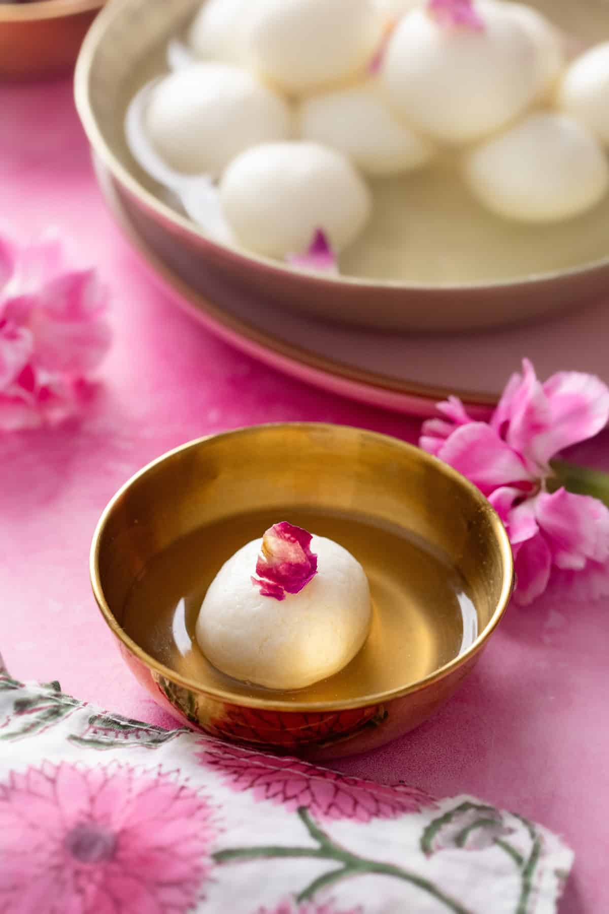 A bowl with a single rasgulla garnished with a pink flower petal, set on a pink surface with flowers. In the background, a larger bowl holds several rasgullas in syrup.