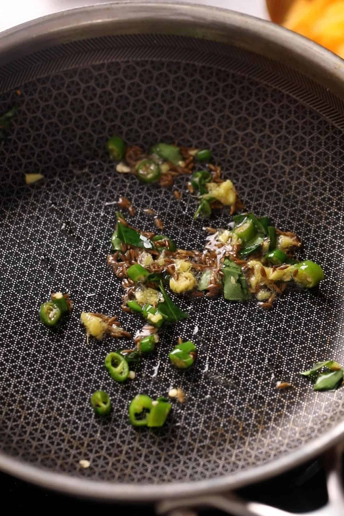 Oil is tempered with cumin, fresh herbs and green chilli in a black pan.