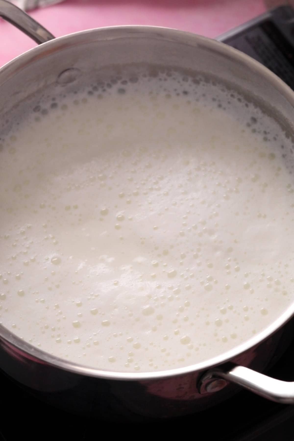 A stainless steel pot filled with boiling milk, with bubbles forming on the surface, sits on a stovetop.