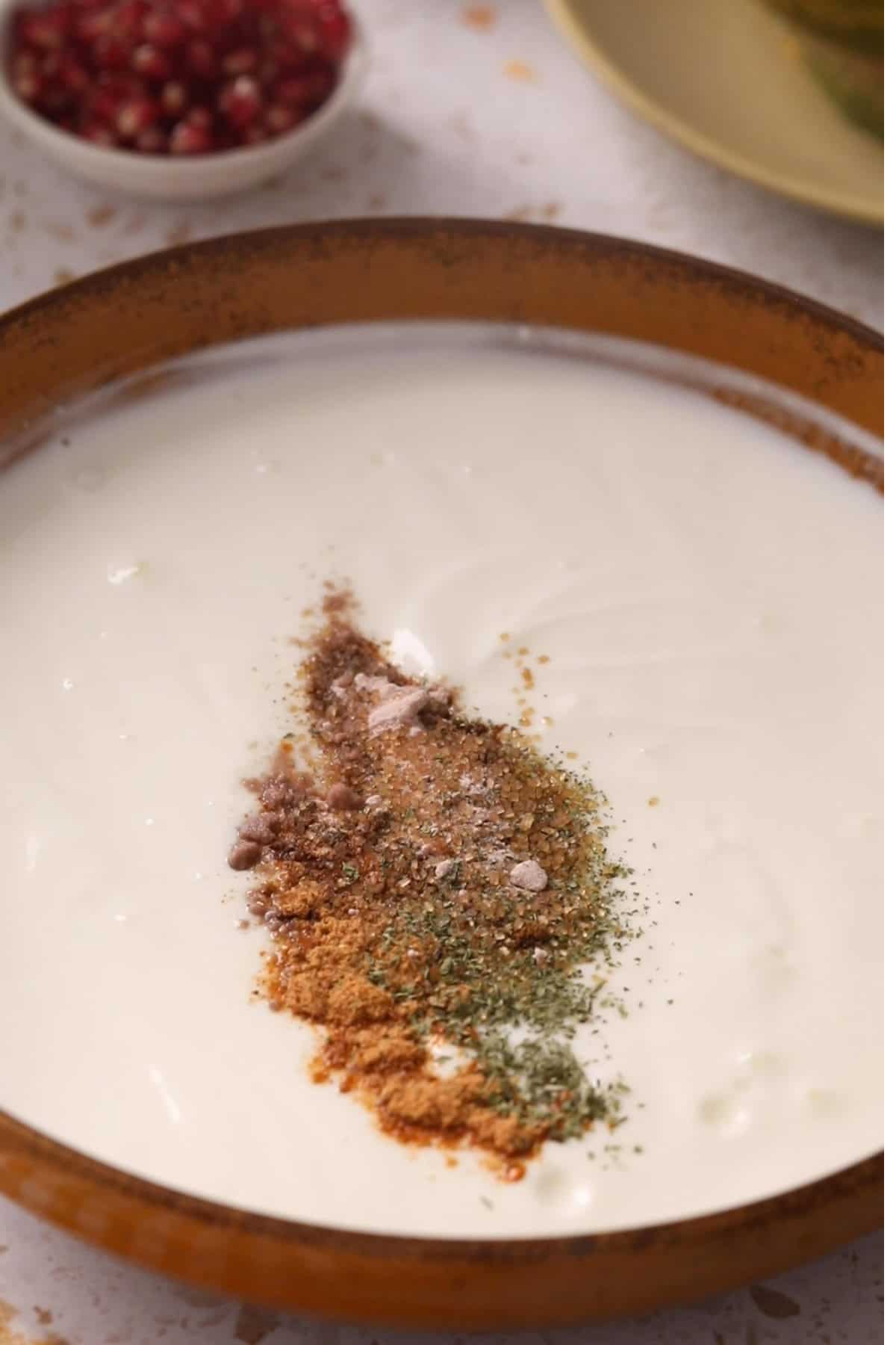 Ground spices are added to whisked yogurt in a brown bowl.