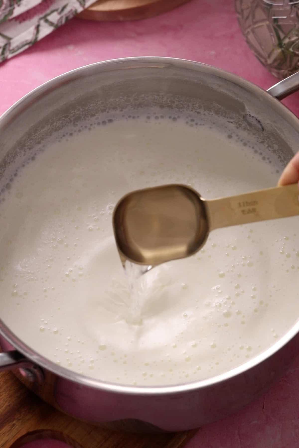 A hand pours a tablespoon of vinegar into a pot of foamy milk.
