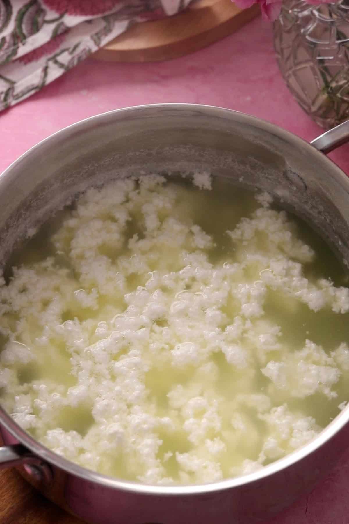 A stainless steel pot filled with curdled milk and yellowish whey, likely in the process of making cheese or paneer, sits on a pink surface.