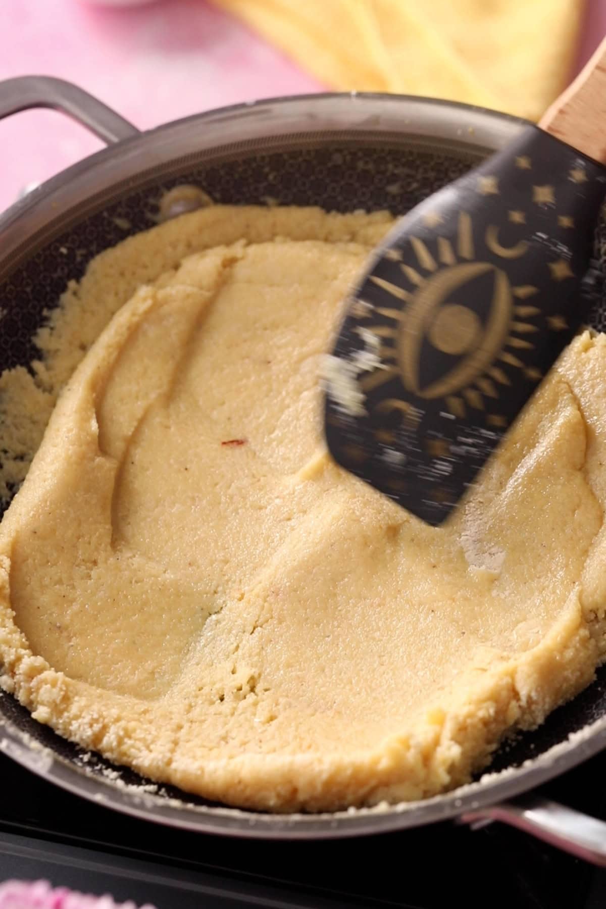 Khoya badam burfi mixture is soft and pilable and being folded with a black spatula.