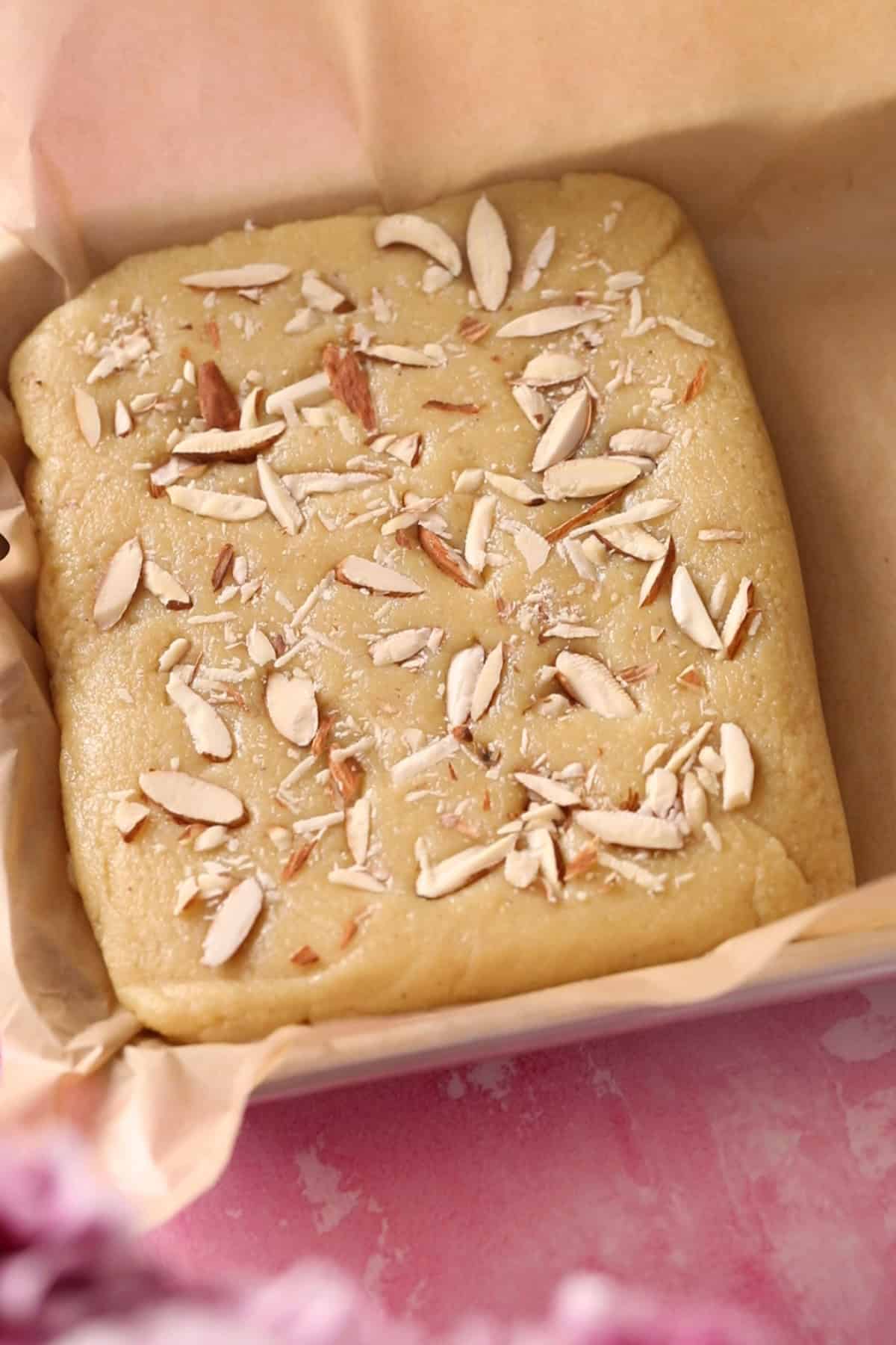 Badam Burfi is set in a brown parchment paper lined tray and sliced alomnds are pressed on top.