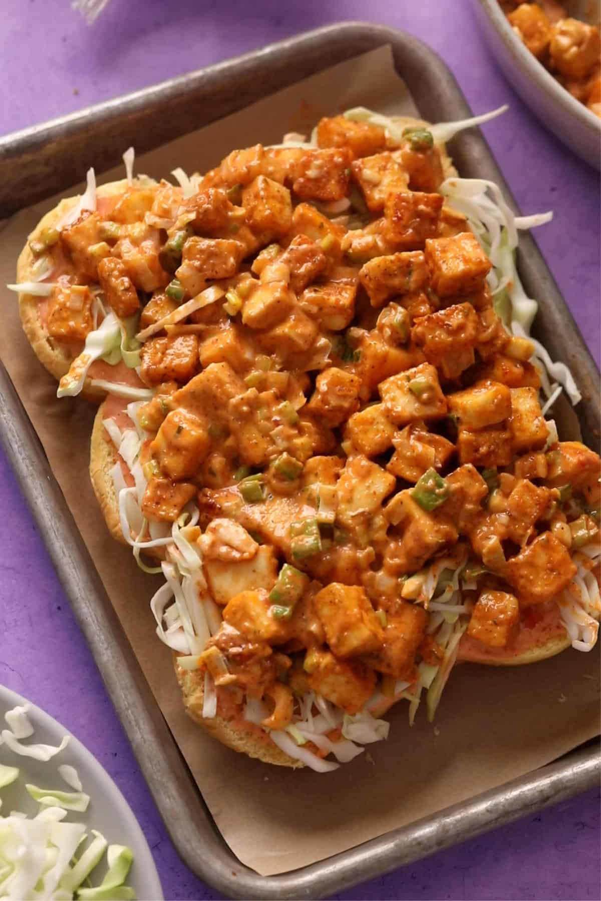 Sliced slider buns on a baking sheet, each topped with creamy bang bang dressing,paneer cubes and, shredded cabbage.
