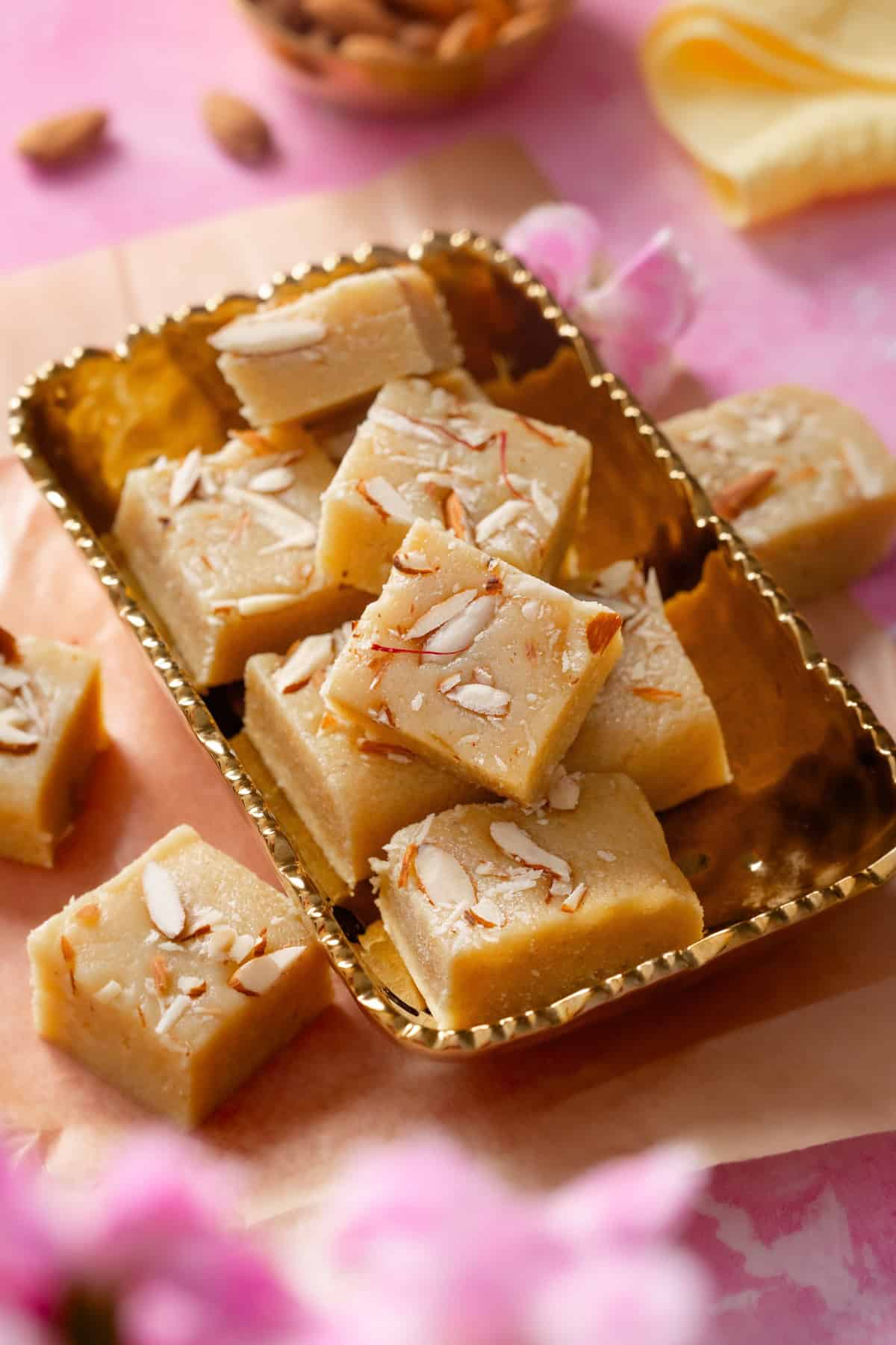 Pieces of khoya badam burfi are placed in a golden rectangular dish with pink flower in background.