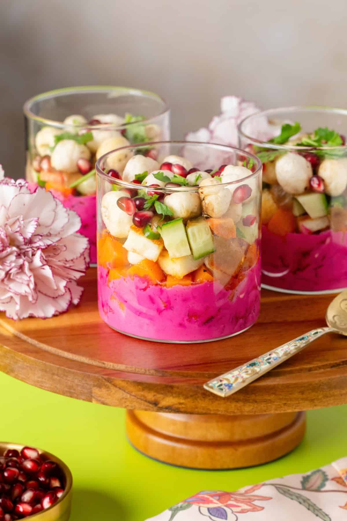 Three chaat cups glass jars with bright pink beet dip, cubed vegetables, pomegranate seeds, and puffed grains, served on a wooden pedestal.