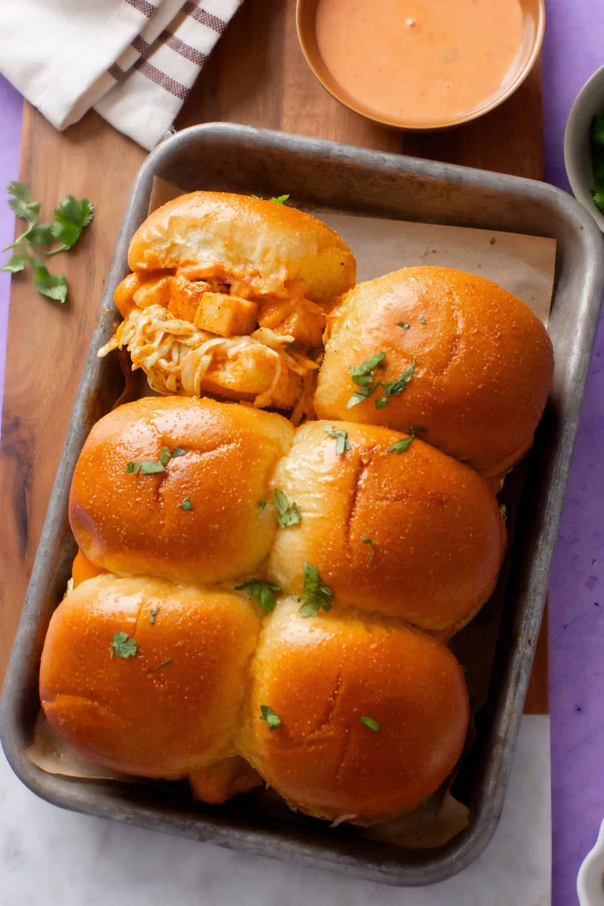 A metal tray of four baked paneer sliders topped with fresh cilantro, served with a side of bang bang sauce.