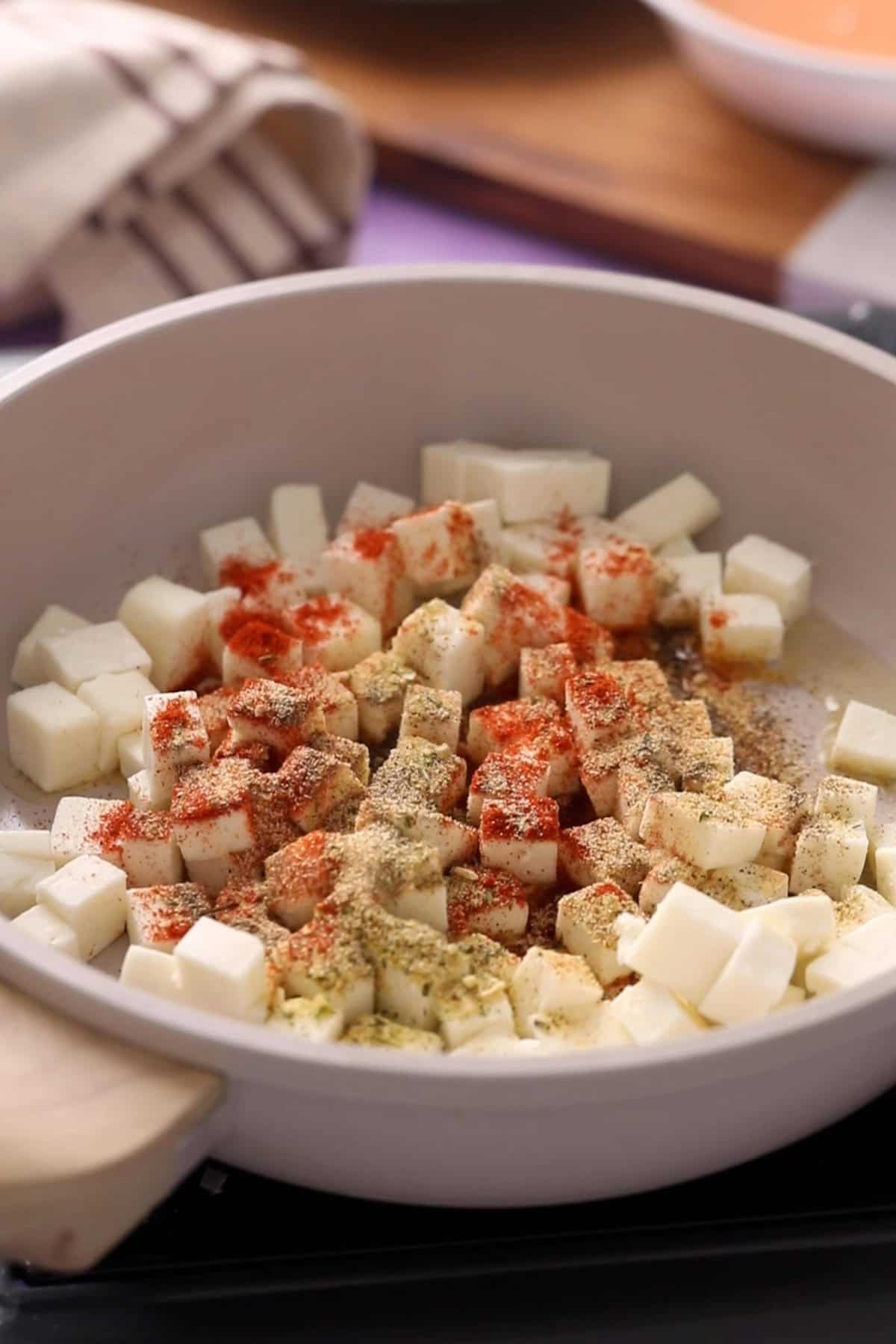 Cubed paneer in a pan, seasoned with red chili powder and dried herbs.