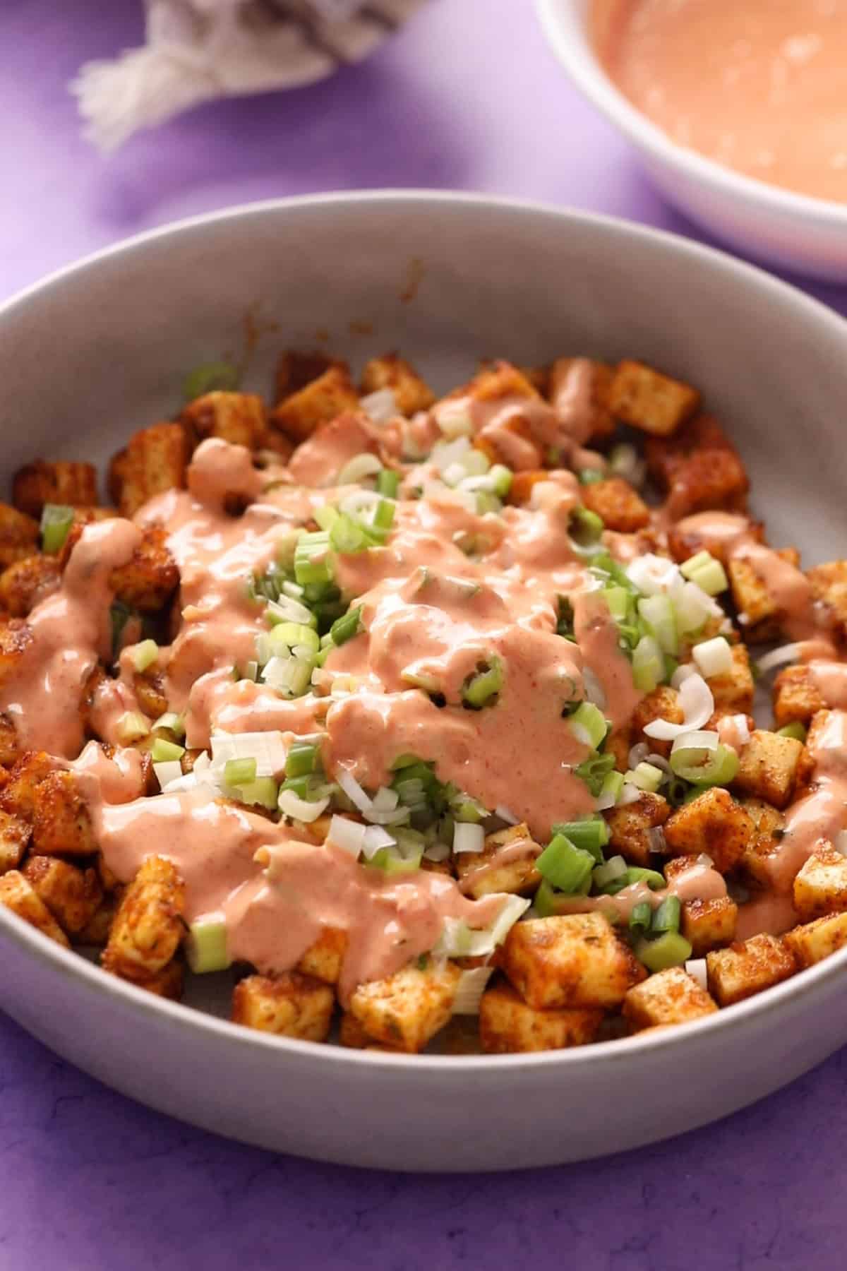 Bowl of cubed, seasoned paneer topped with chopped green onions and a creamy bang bang sauce.